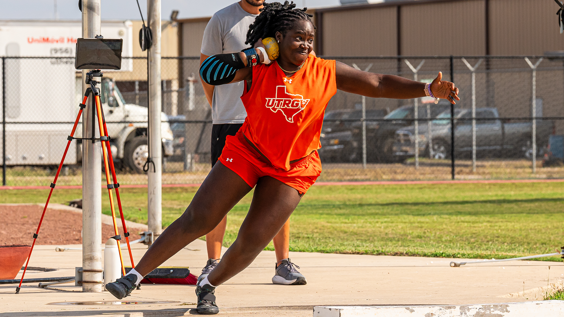 Photo of Efe Latham in an orange UTRGV track & field shirt and orange shorts mid-turn as she's shot putting in the throwing ring at UTRGV Soccer and Track & Field Complex.
