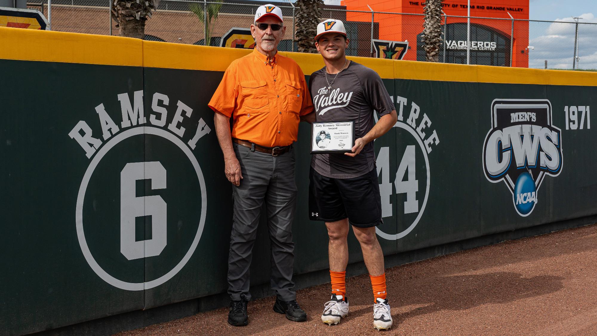 Hank Warren, holding the Jody Ramsey Memorial Award, standing with Kenneth Ramsey next to the Ramsey 6 on the left field wall at UTRGV Baseball Stadium