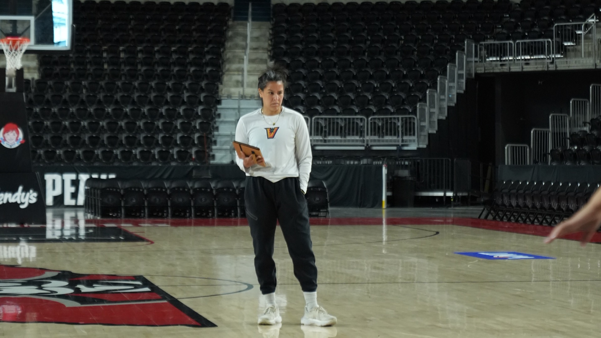 Image shows UTRGV WBB assistant coach Victoria Pena in a white shirt and black pants holding an iPad as she oversees a drill at practice in the Austin Peay gym.