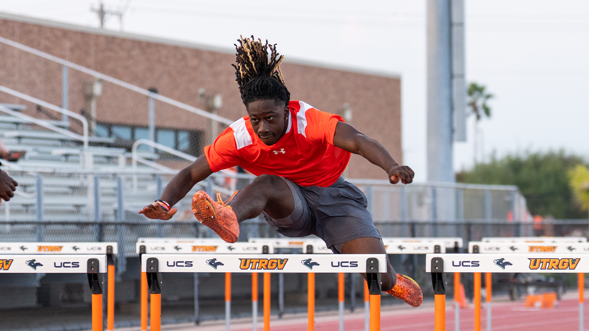 JaDan Keiler in an orange shirt is jumping over a hurdle that says UTRGV on the track.
