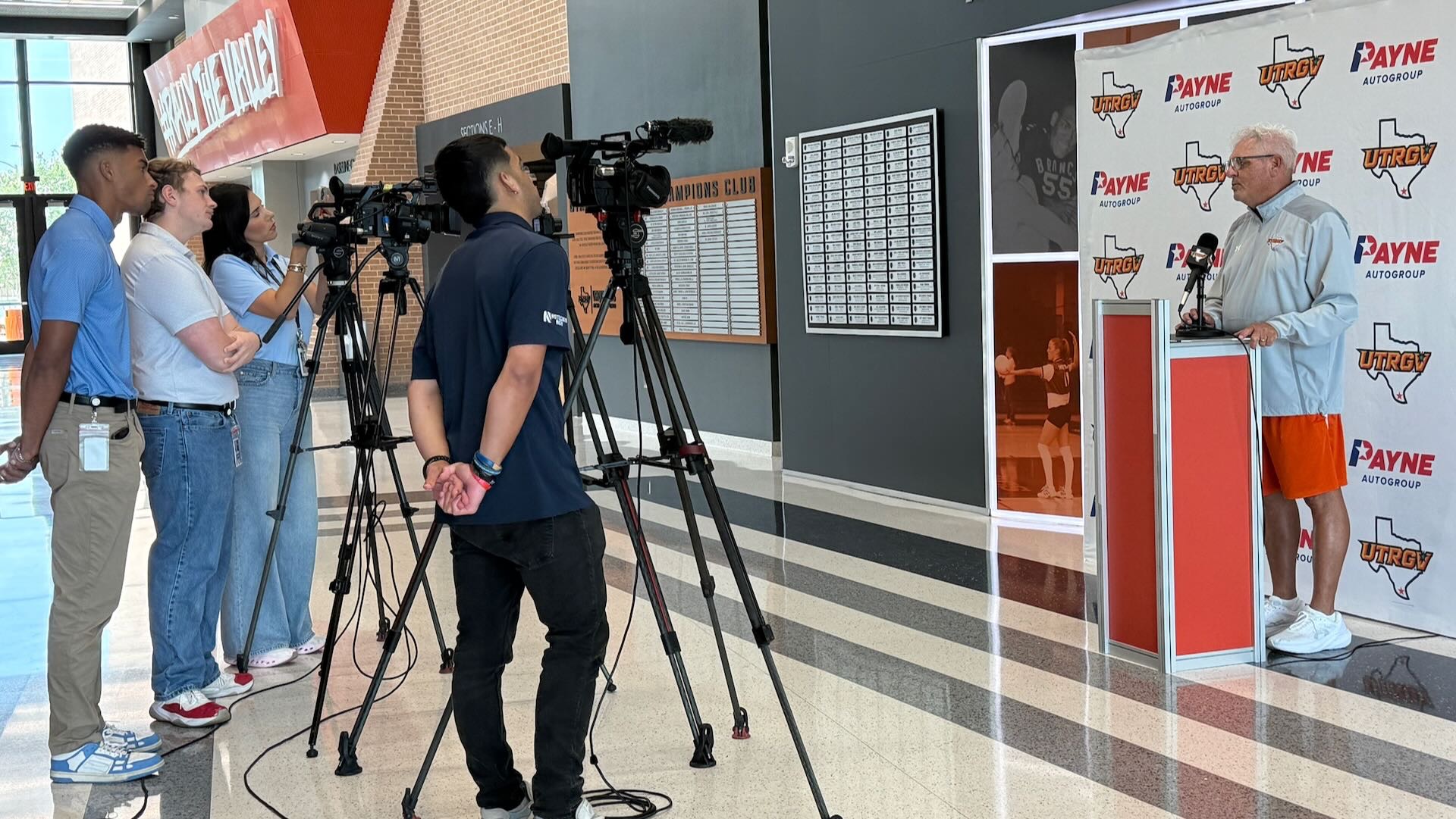 Photo shows head coach Lane Lord standing at a podium in front of a Payne and UTRGV white backdrop. Four media members are shown standing in front of their cameras on tripods watching Lord speak.