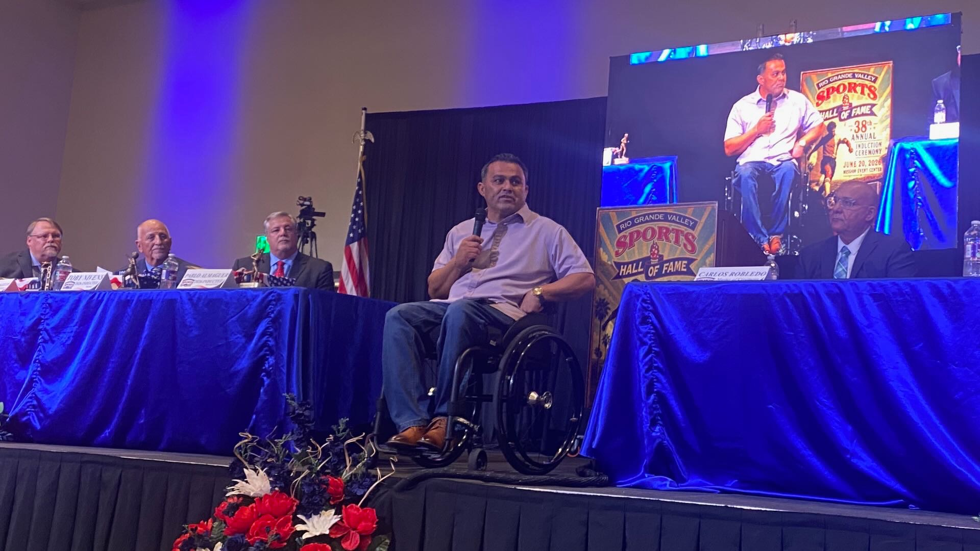 Image shows Pablo Almaguer on stage delivering his speech after being inducted to the RGV Sports Hall of Fame. Other inductees are shown sitting at tables on stage and watching Almaguer speak.