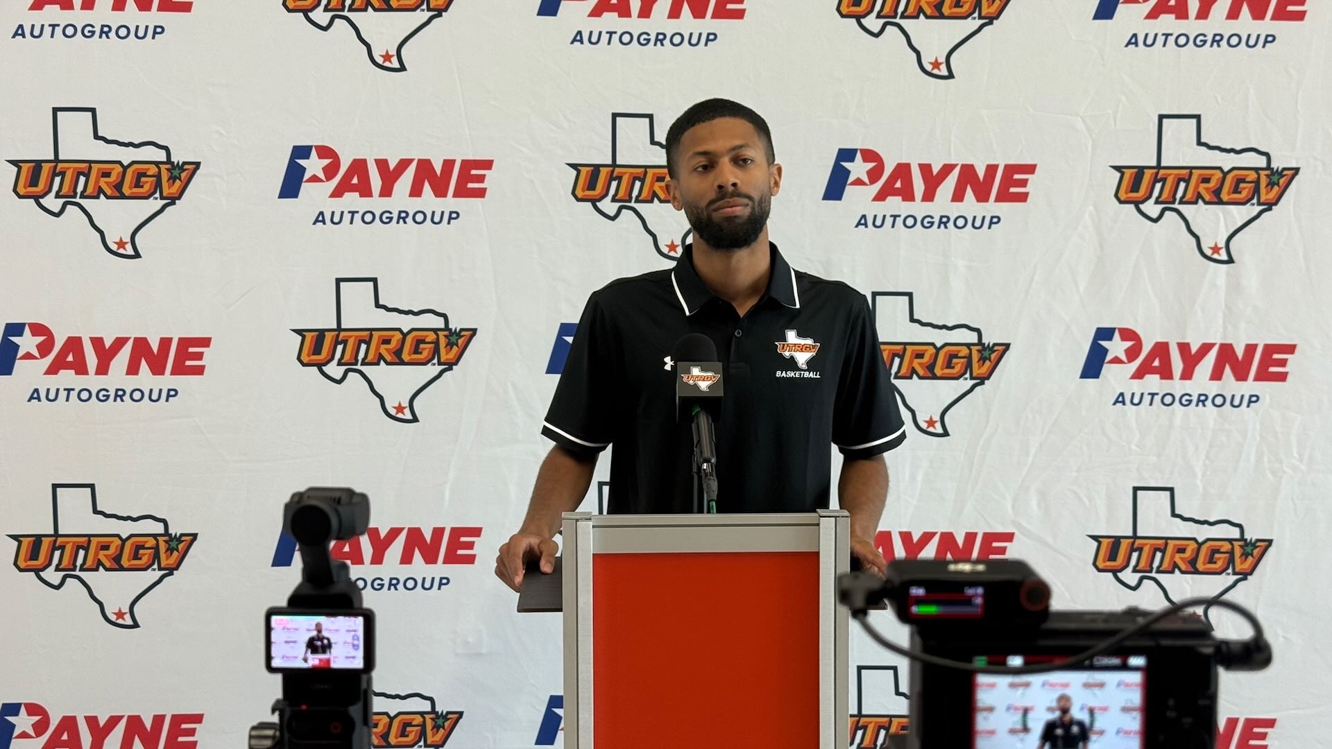 UTRGV men's basketball DOBO John Jones stands at a podium in a black polo in front of a white backdrop with alternating Payne auto group logos and UTRGV over Texas logos.