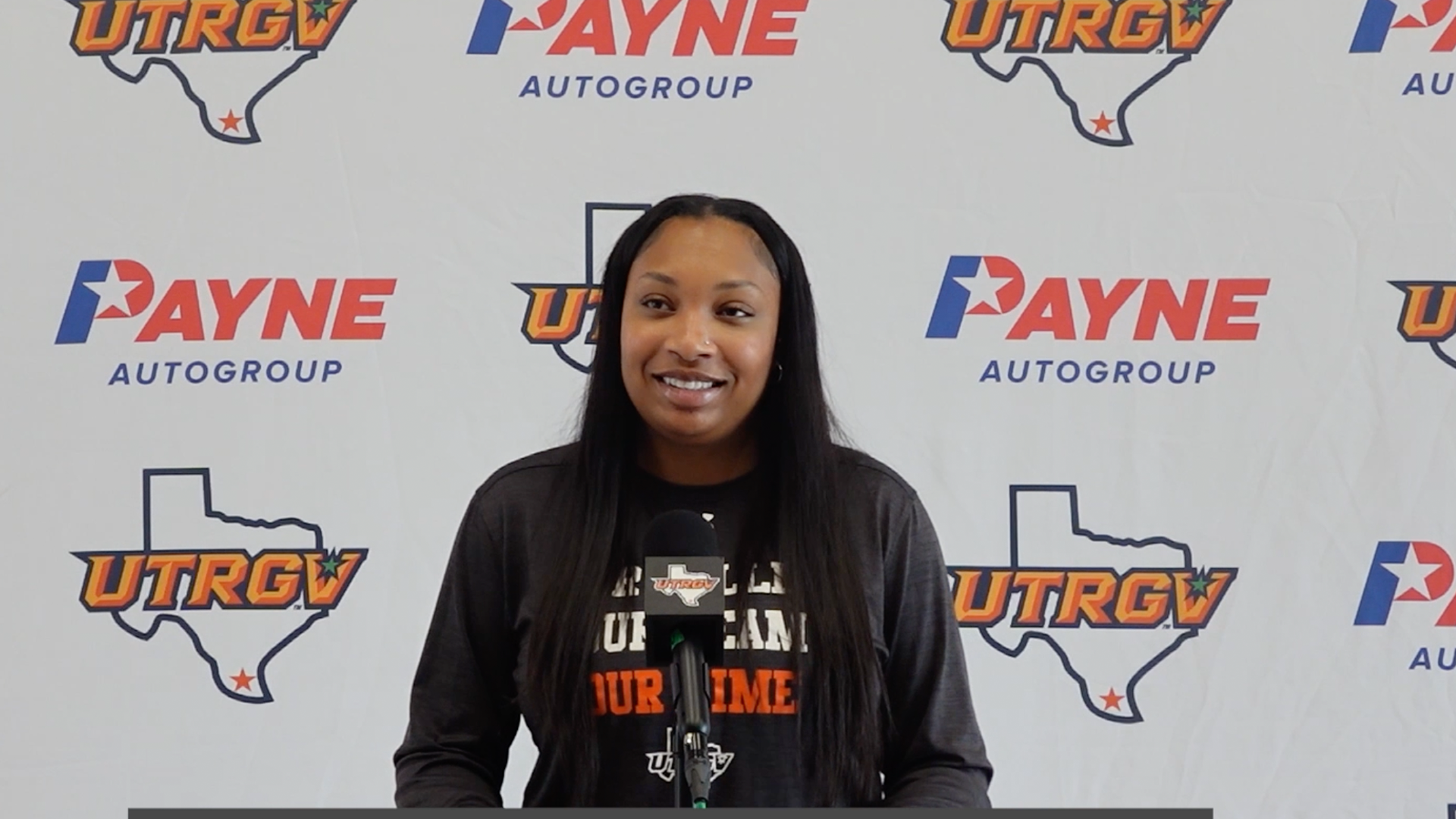 Image shows UTRGV women's basketball assistant coach Jurnee President standing at a podium in a gray long-sleeved shirt in front of a white backdrop with alternating Payne auto group logos and UTRGV over Texas logos.