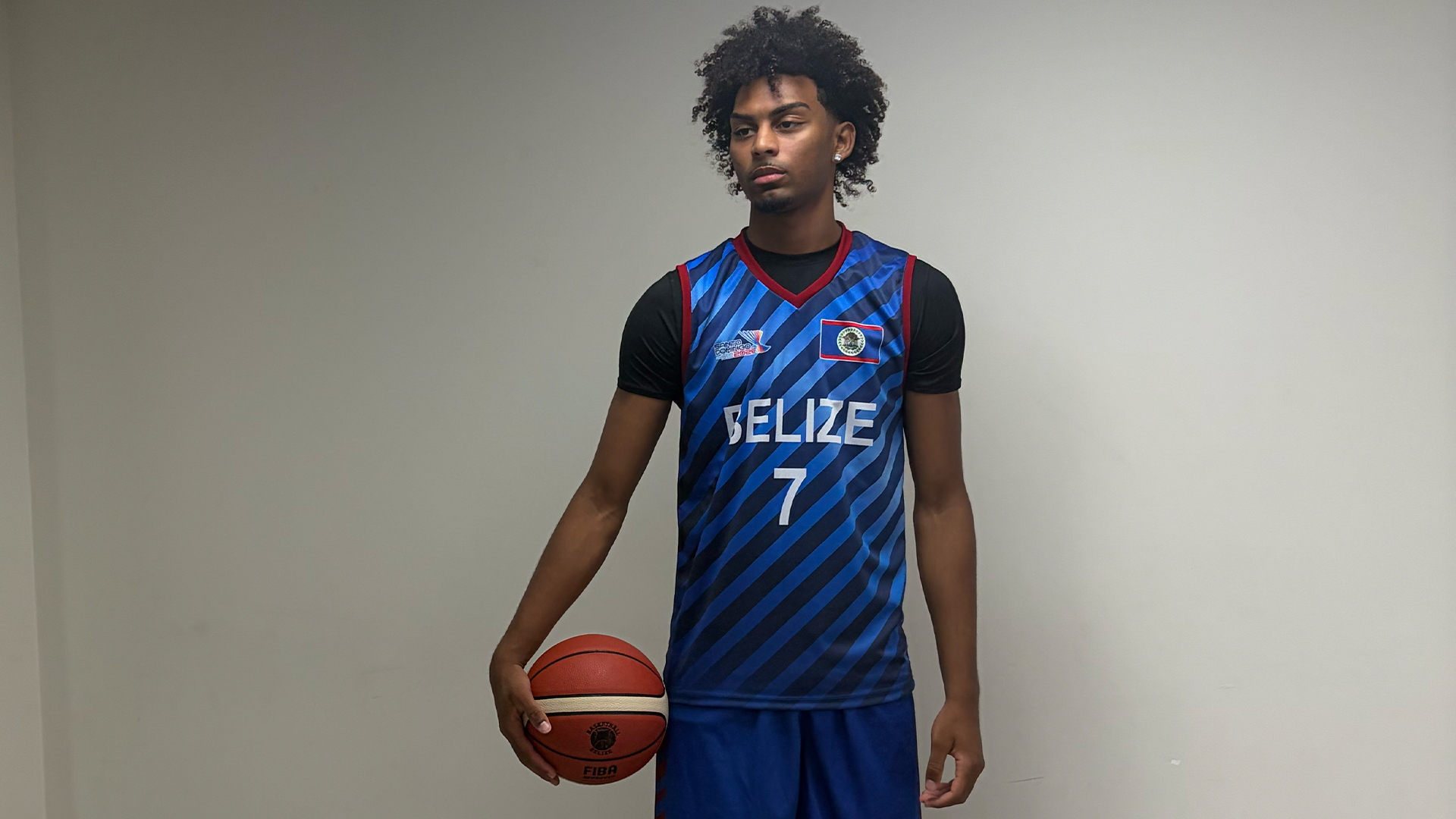 Image shows Ian Parham Jr. wearing a blue basketball uniform with Belize across the chest and the number 7 while holding a basketball posing for a photo in front of a white wall.