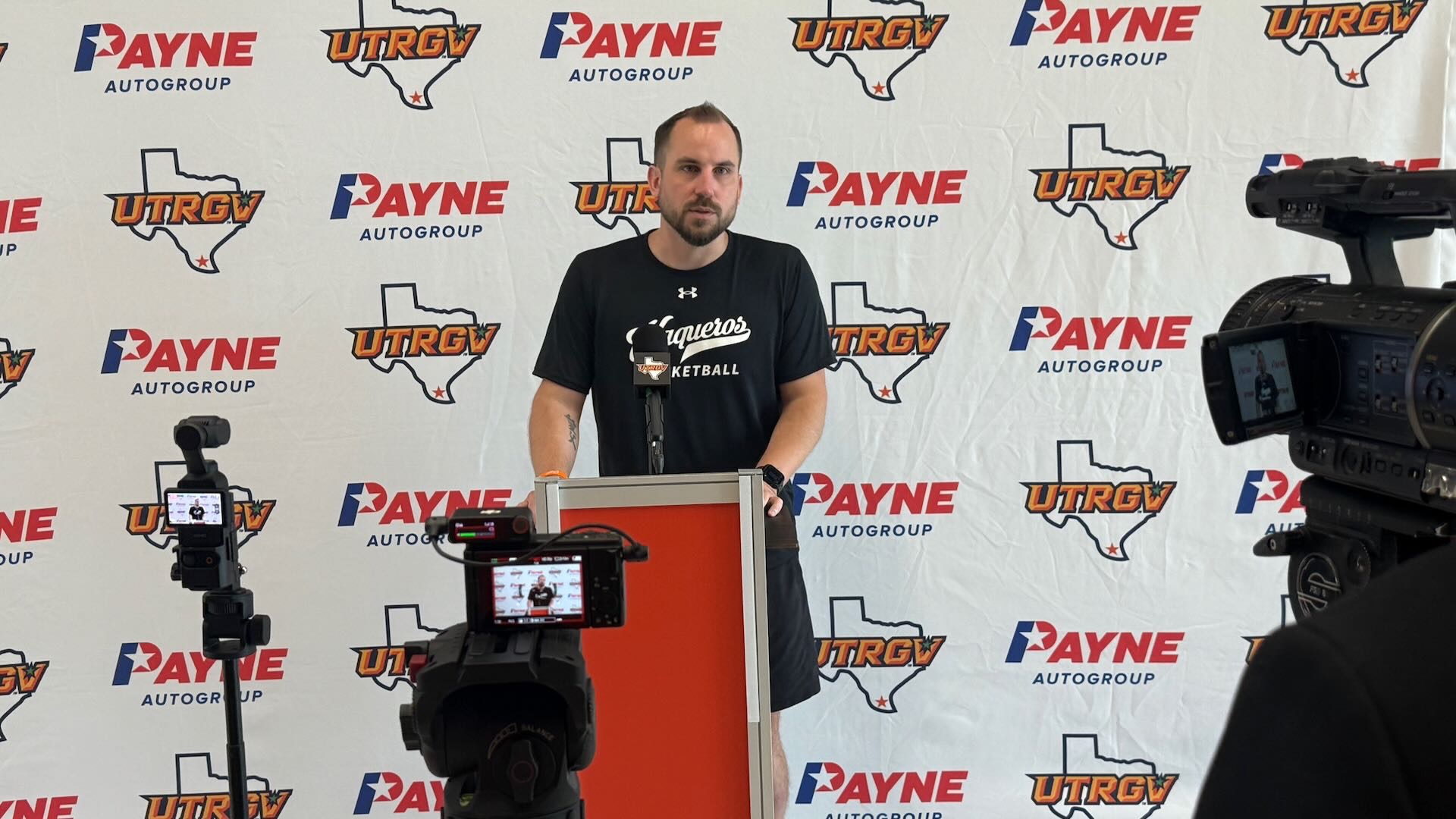 Image shows UTRGV men's basketball head coach Brandon Chambers standing at a podium with three cameras pointed at him as he addresses media in front of a white backdrop with alternating Payne auto group and UTRGV over Texas logos.