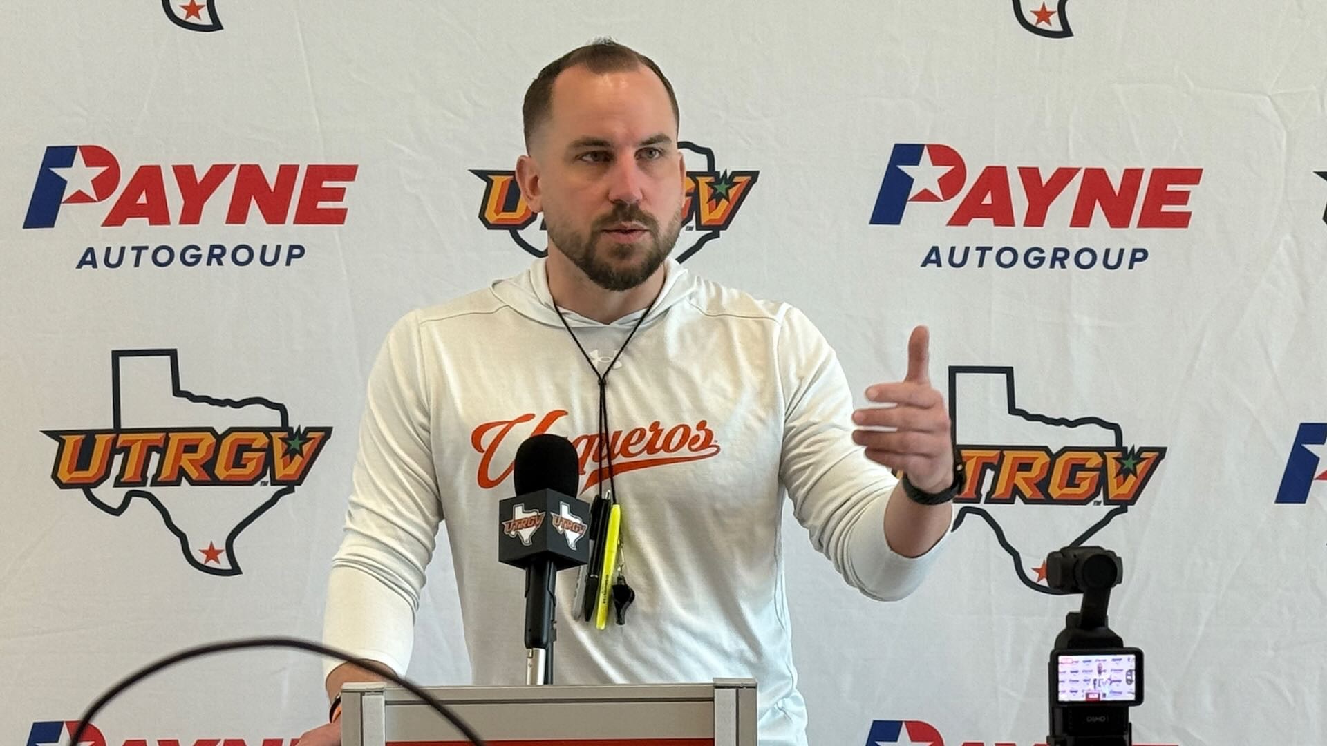 Image shows UTRGV men's basketball head coach Brandon Chambers addressing media behind a podium in front of a white backdrop with alternating Payne auto group logos and UTRGV over Texas logos.