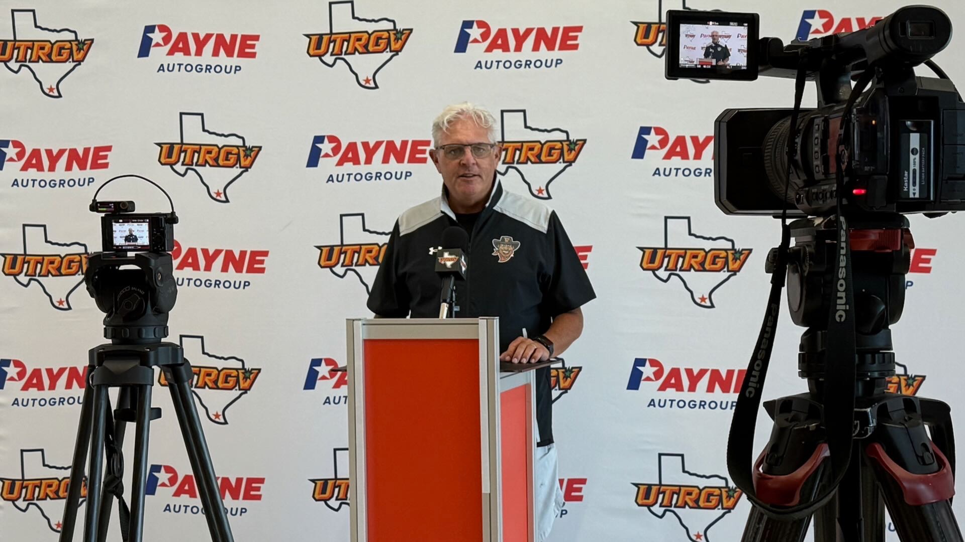 Image shows head coach Lane Lord standing at a podium in front of a white backdrop with alternating UTRGV over Texas logos and Payne auto group logos with two cameras on tripods pointing at him recording his weekly press conference.