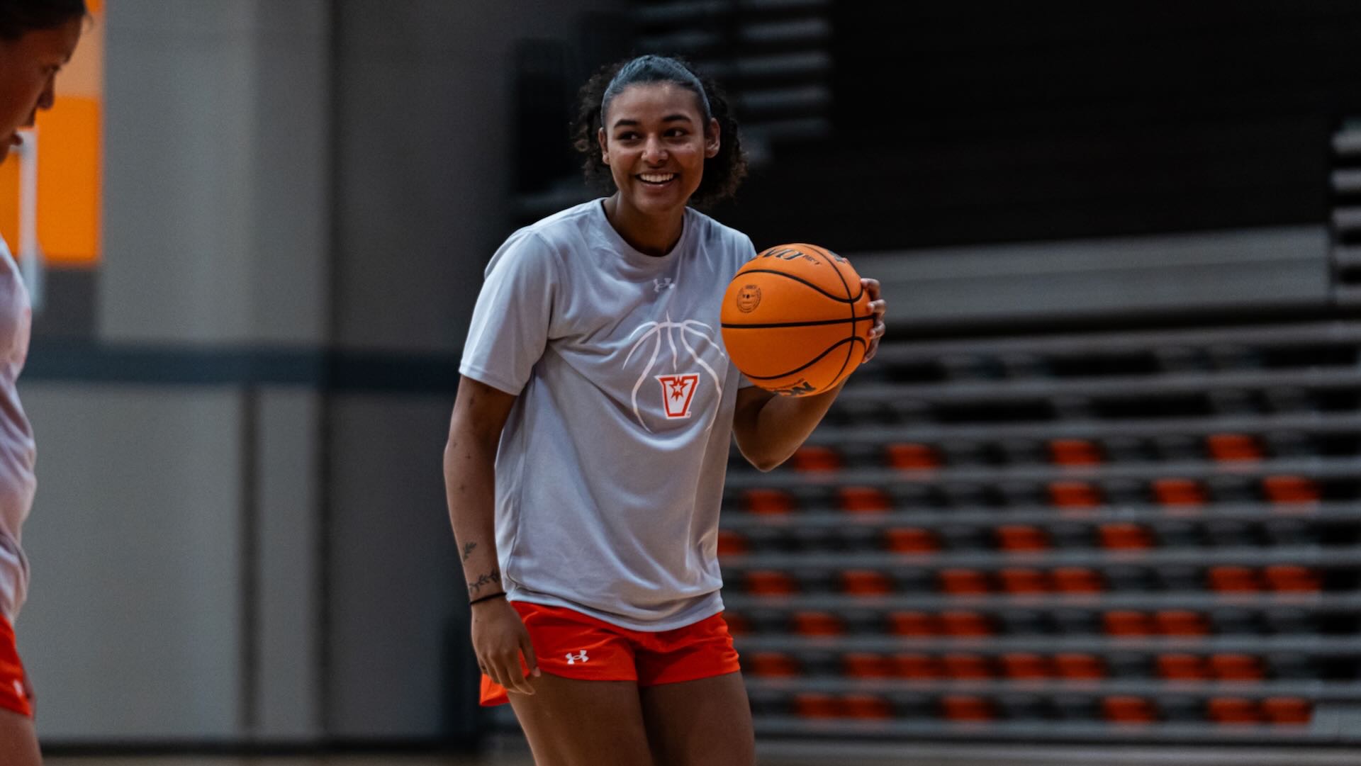 Image shows women's basketball student-athlete Trinity Bane in a gray practice shirt and orange shorts smiling while dribbling a basketball at the UTRGV Fieldhouse.
