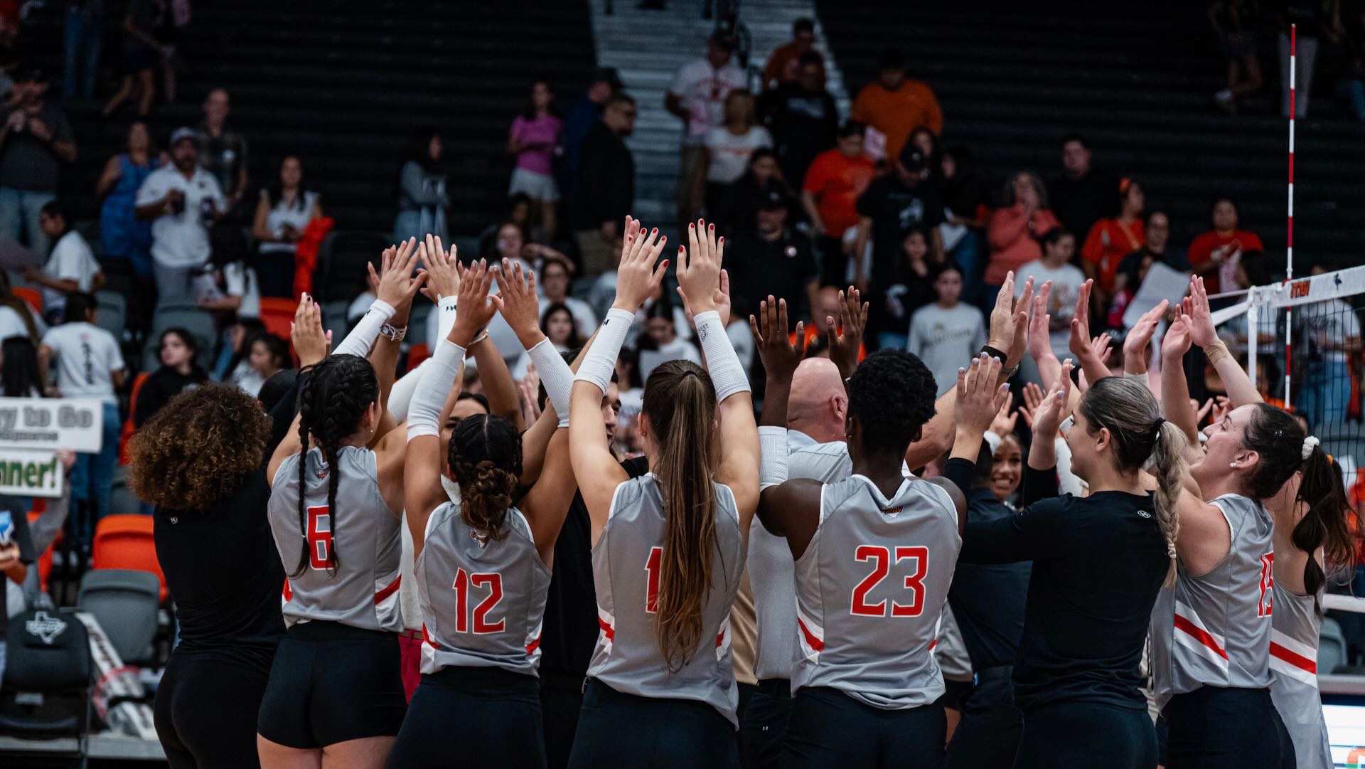 Image shows the UTRGV volleyball team holding their hands up celebrating in a huddle.