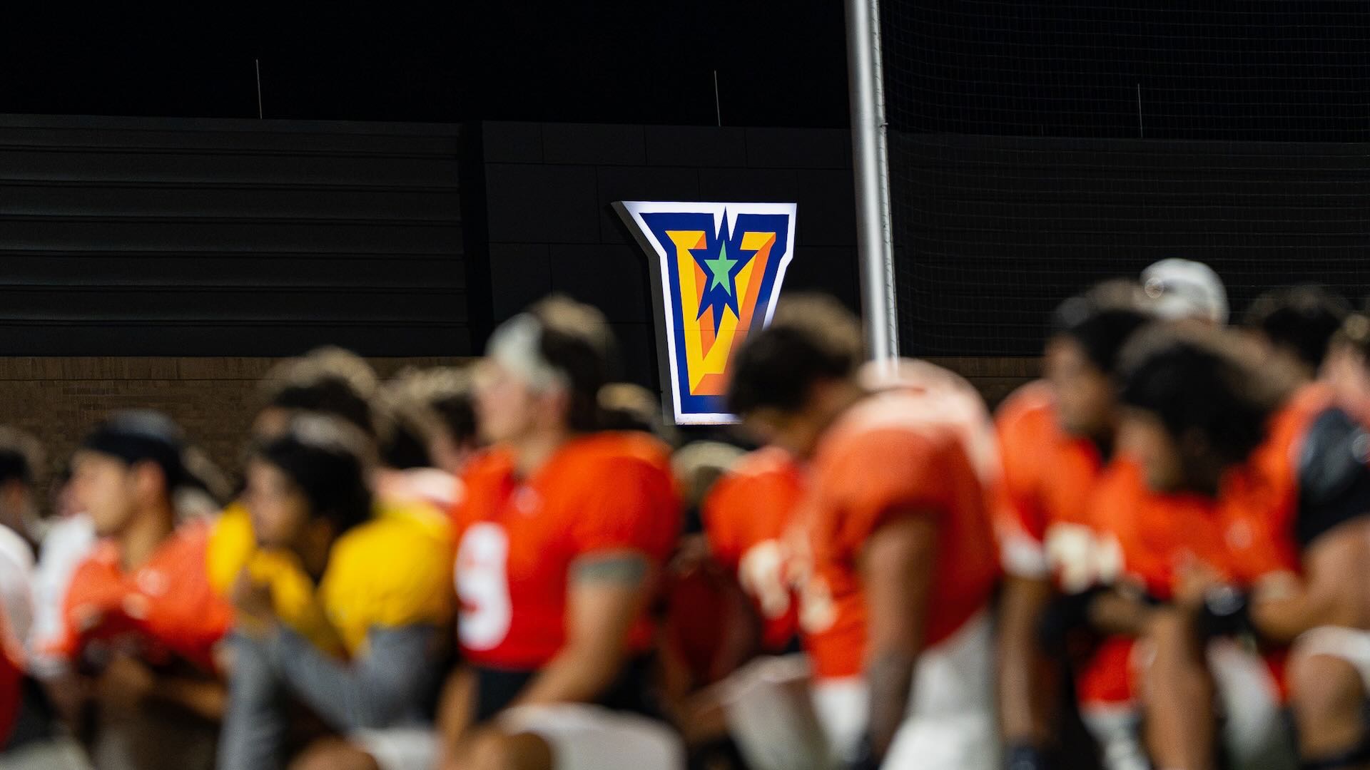 Image focuses on a lit V alternate UTRGV logo with football players kneeling out of focus in the foreground.