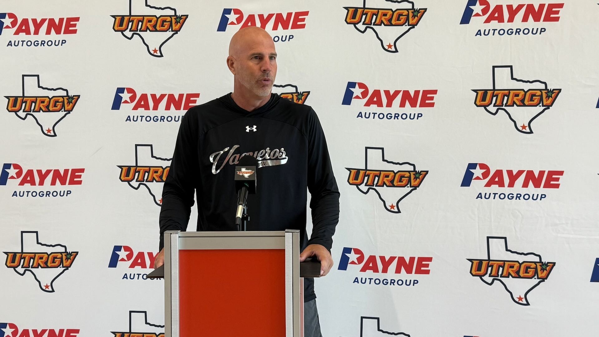 Image shows UTRGV Volleyball head coach Todd Lowery in a black long-sleeve shirt with Vaqueros script across the chest standing at a podium in front of a white backdrop with alternating Payne auto group and UTRGV over Texas logos.