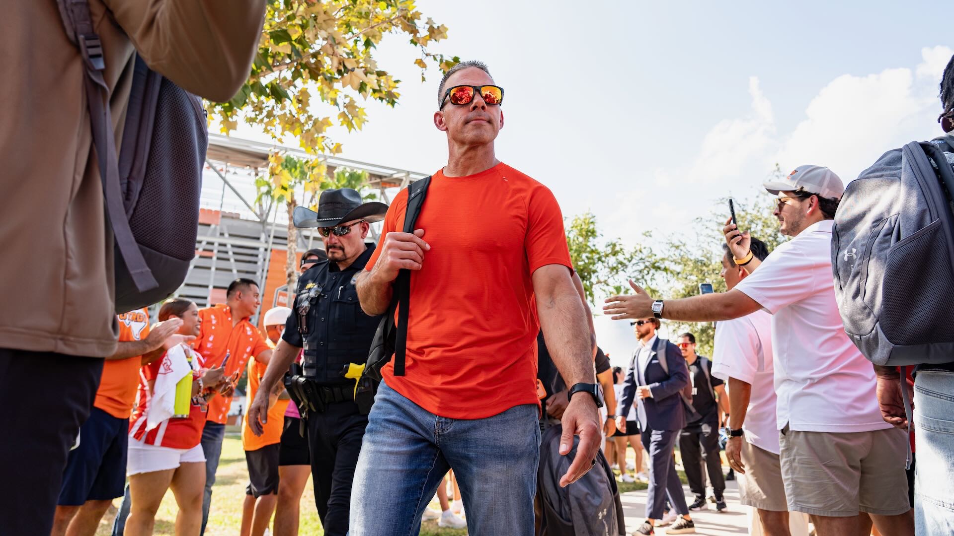 Image of a man in an orange shirt and jeans and sunglasses walking into the stadium.