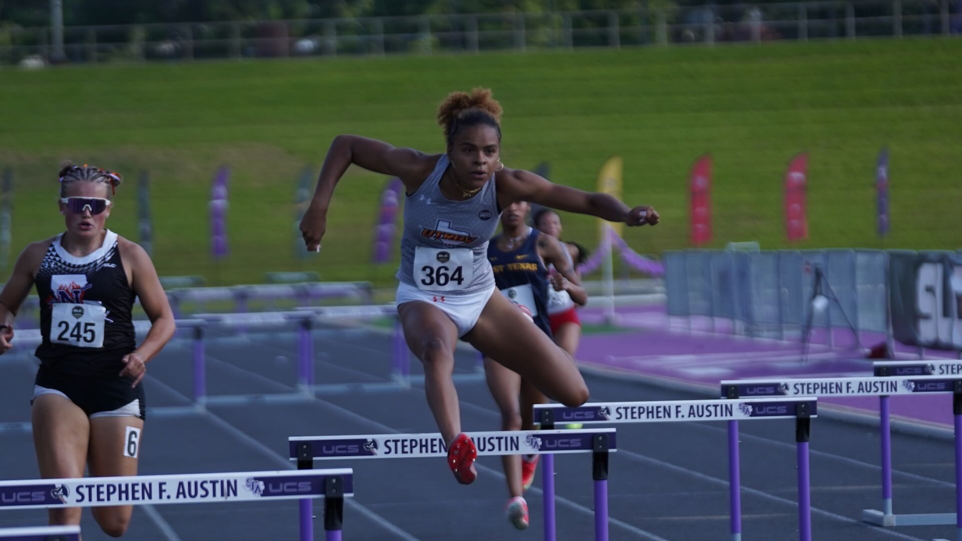 Image shows UTRGV track & field student-athlete Talyna Ligier in a gray UTRGV uniform jumping over a hurdle on the track.