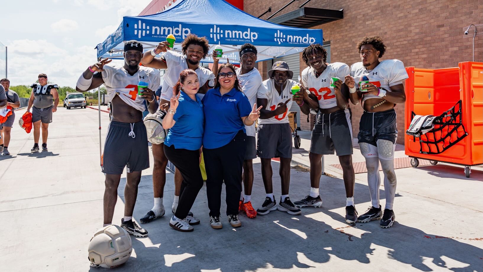 Members of the UTRGV football team pose with two workers with NMDP in front of a tent with snow cones after practice