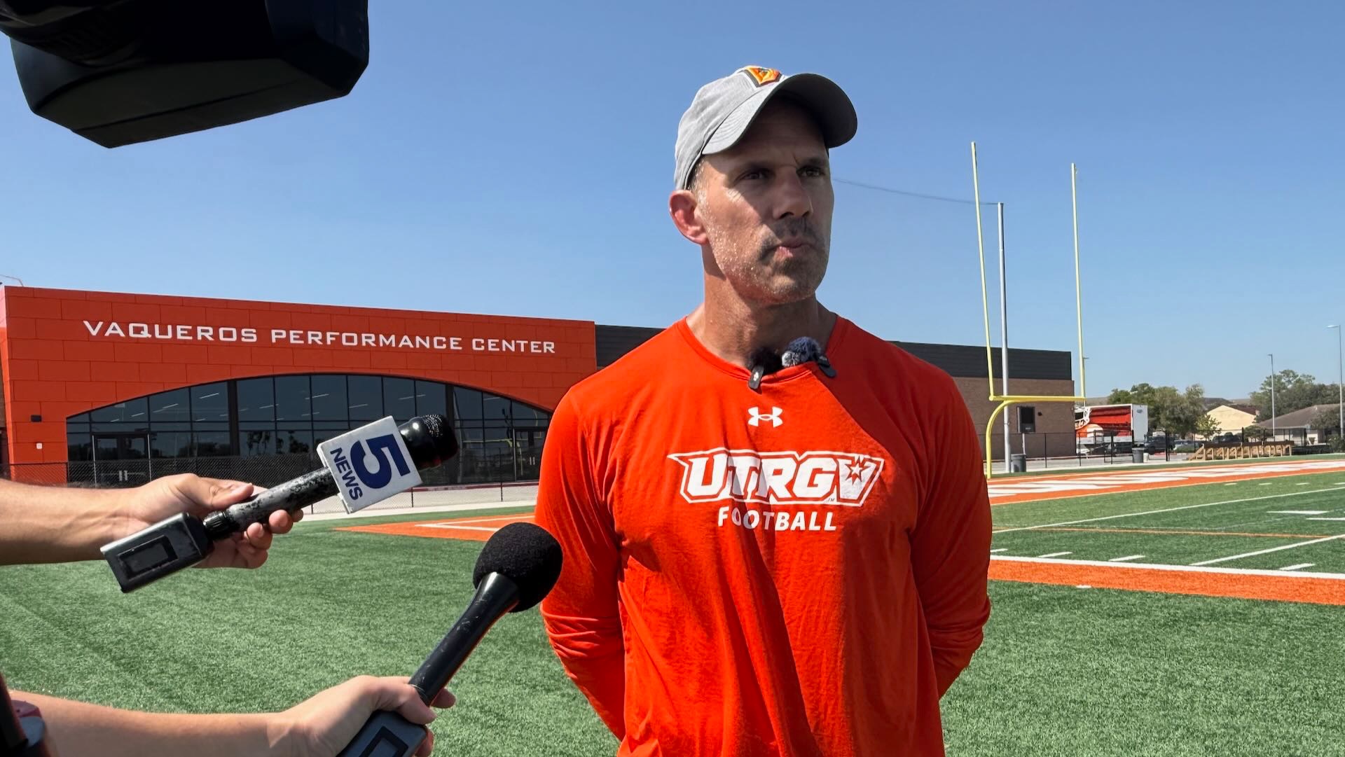 Image shows a man in an orange long-sleeve shirt with a gray cap standing in front of the Vaqueros Performance Center with microphones pointed toward him during a press conference on a football field.