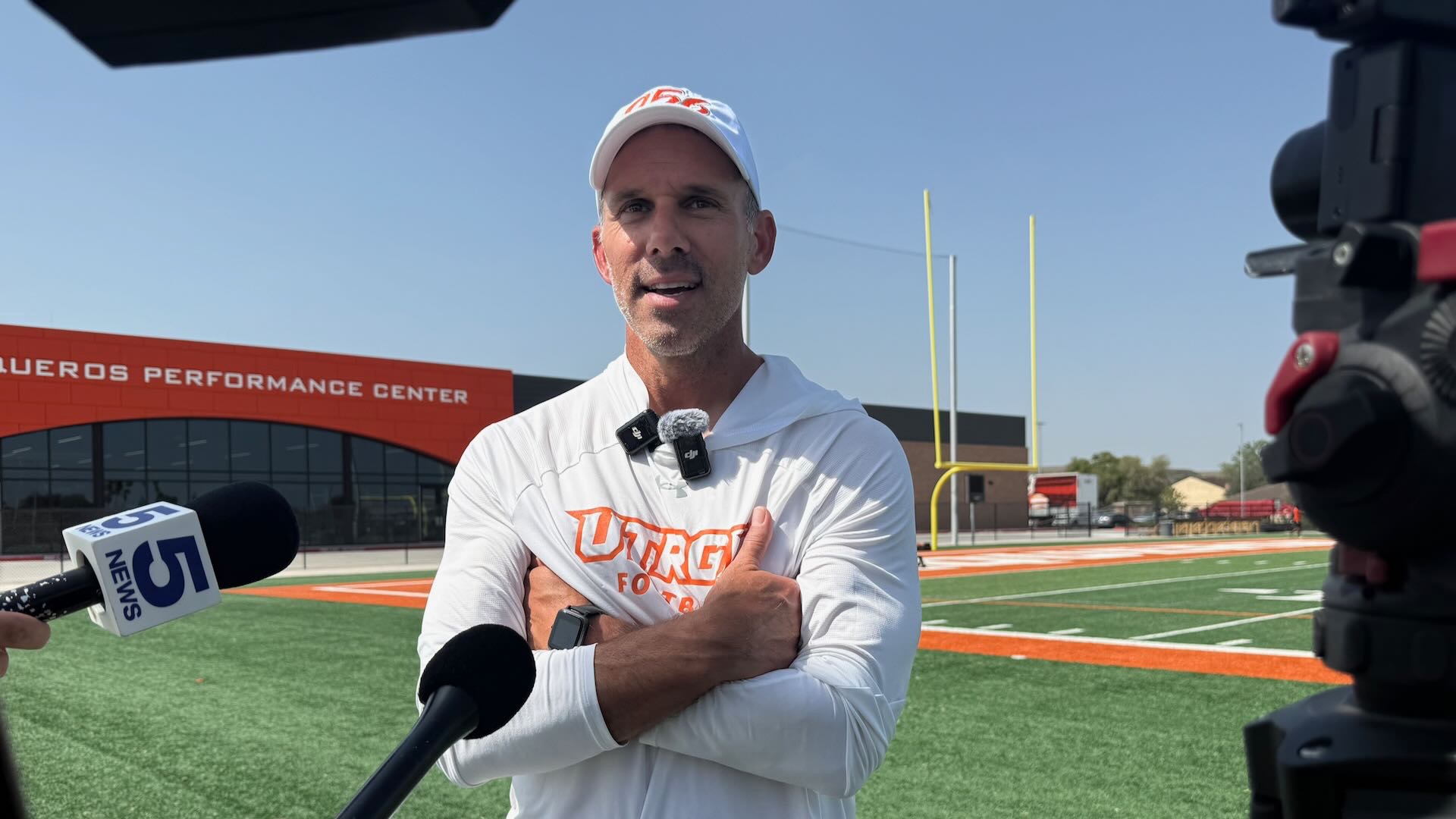 Image shows UTRGV Football head coach Travis Bush standing in a white shirt with mics on his collar in front of the Vaqueros Performance Center addressing media.