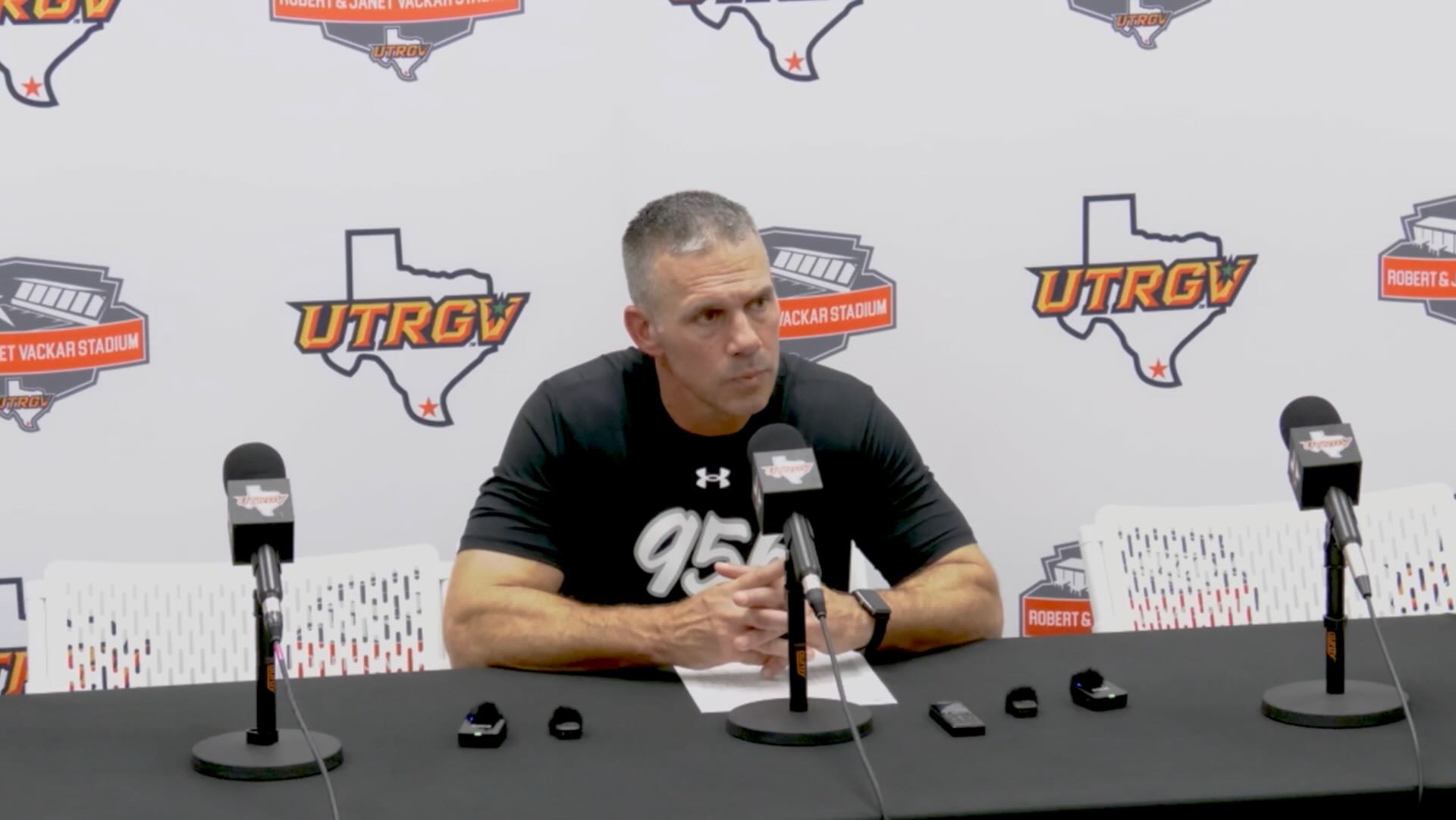 Image shows head UTRGV football coach Travis Bush sitting at a table in front of three mics and some mini microphones addressing media with a white backdrop with alternating Robert & Janet Vackar Stadium and UTRGV Athletics logos.