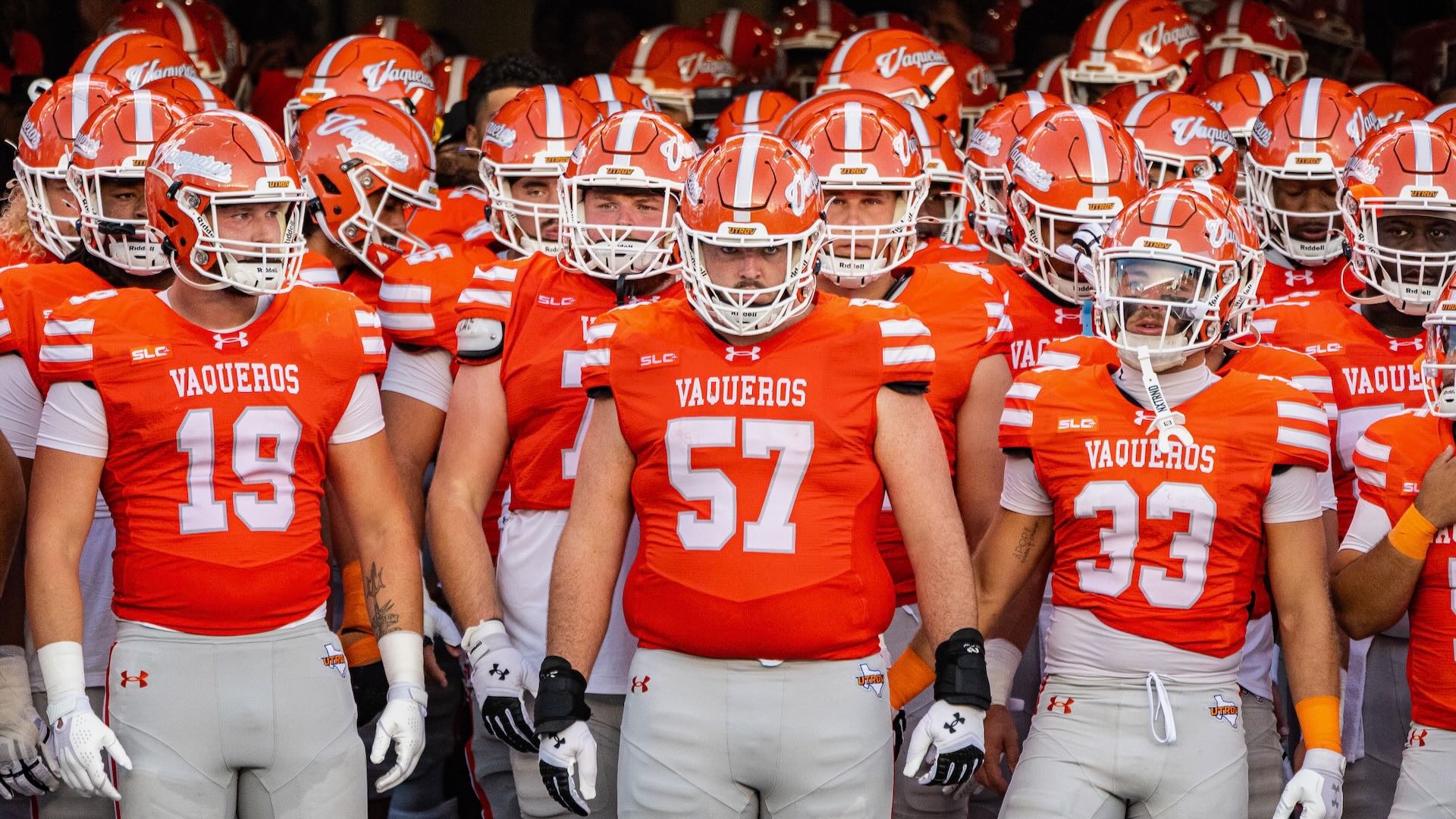 Football players are shown wearing orange uniforms and helmets standing in the tunnel at Robert & Janet Vackar Stadium.
