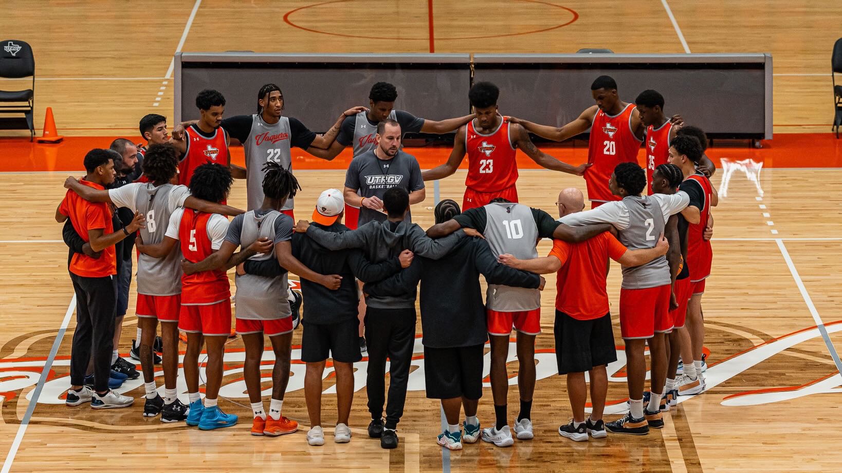 Image shows basketball players huddled together with arms across each other's shoulders looking at a coach standing in the center of the huddle addressing the team in a gym.