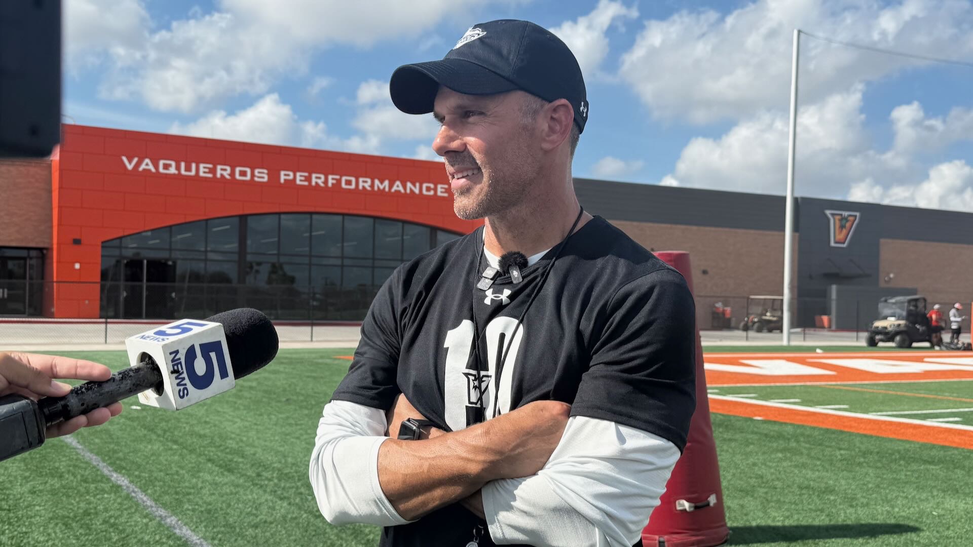 Image shows head football coach Travis Bush in a black shirt and black cap smiling with his arms crossed standing in front of the Vaqueros Performance Center.