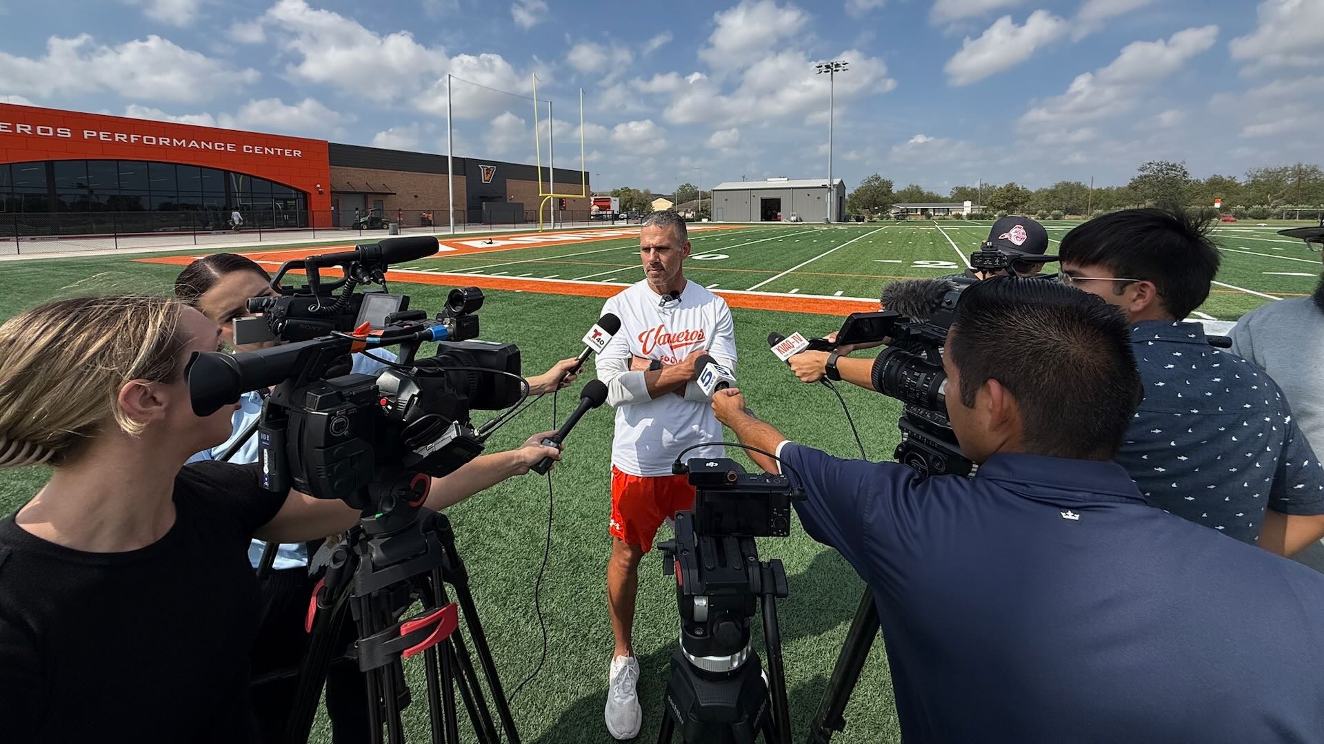 Image shows head coach Travis Bush in a white shirt standing on a field with cameras and microphones pointing at him.