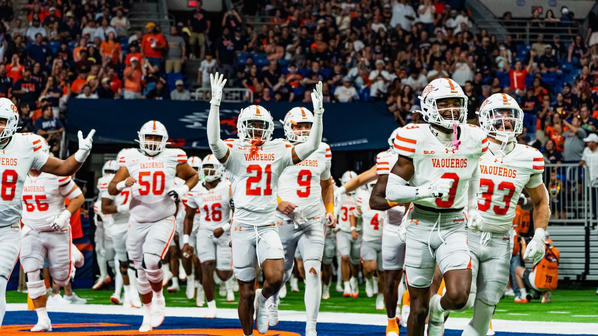 Image shows UTRGV football players in white uniform pants, tops and helmets running onto the field before the game.