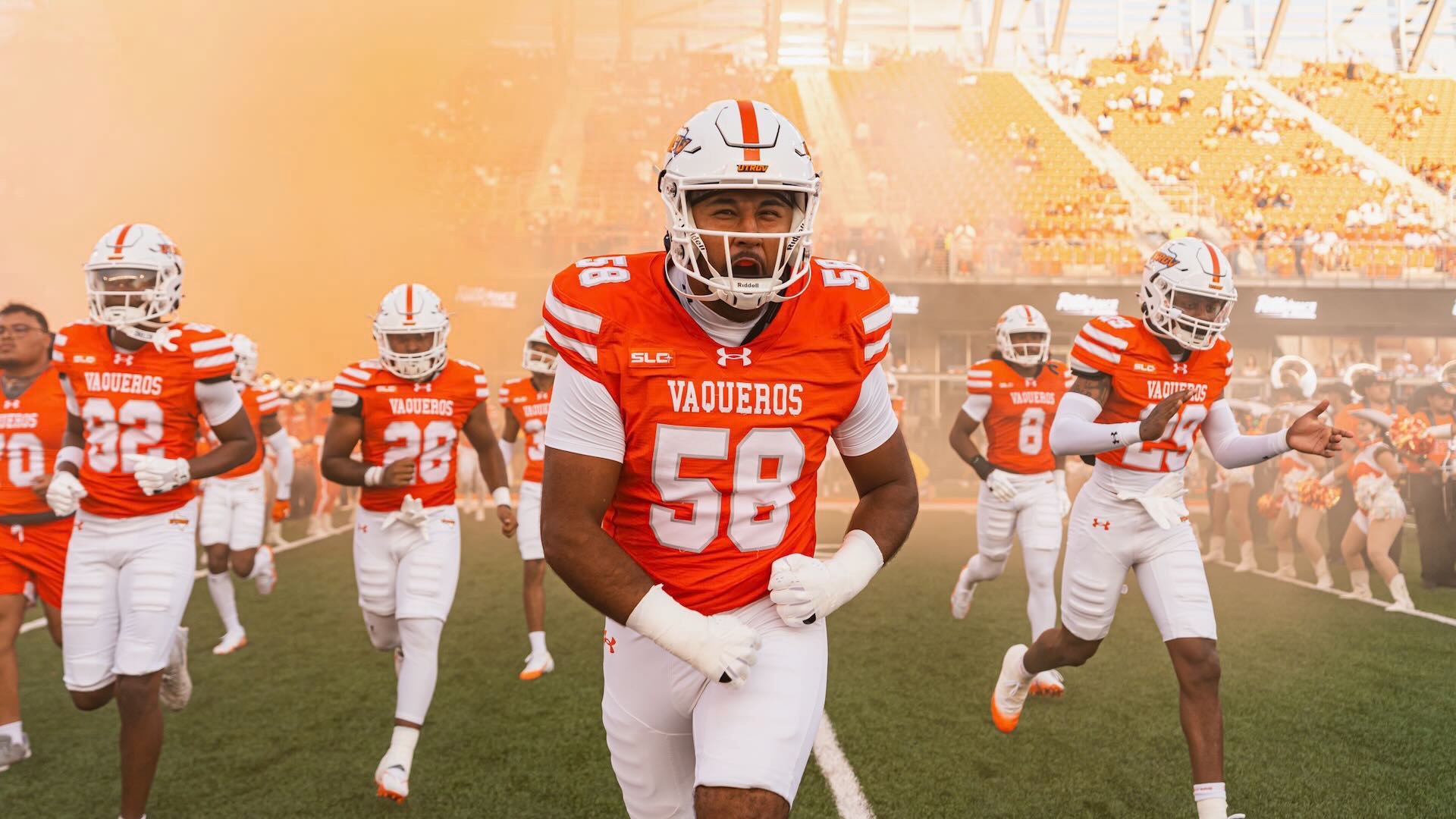 Image shows a football player in a white helmet and orange uniform running onto the field with orange smoke surrounding him.