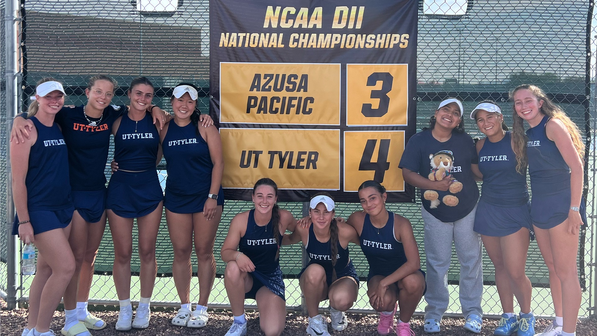 UT Tyler Women's Tennis poses in front of the scoreboard after beating Azusa Pacific 4-3 in the NCAA Round of 16