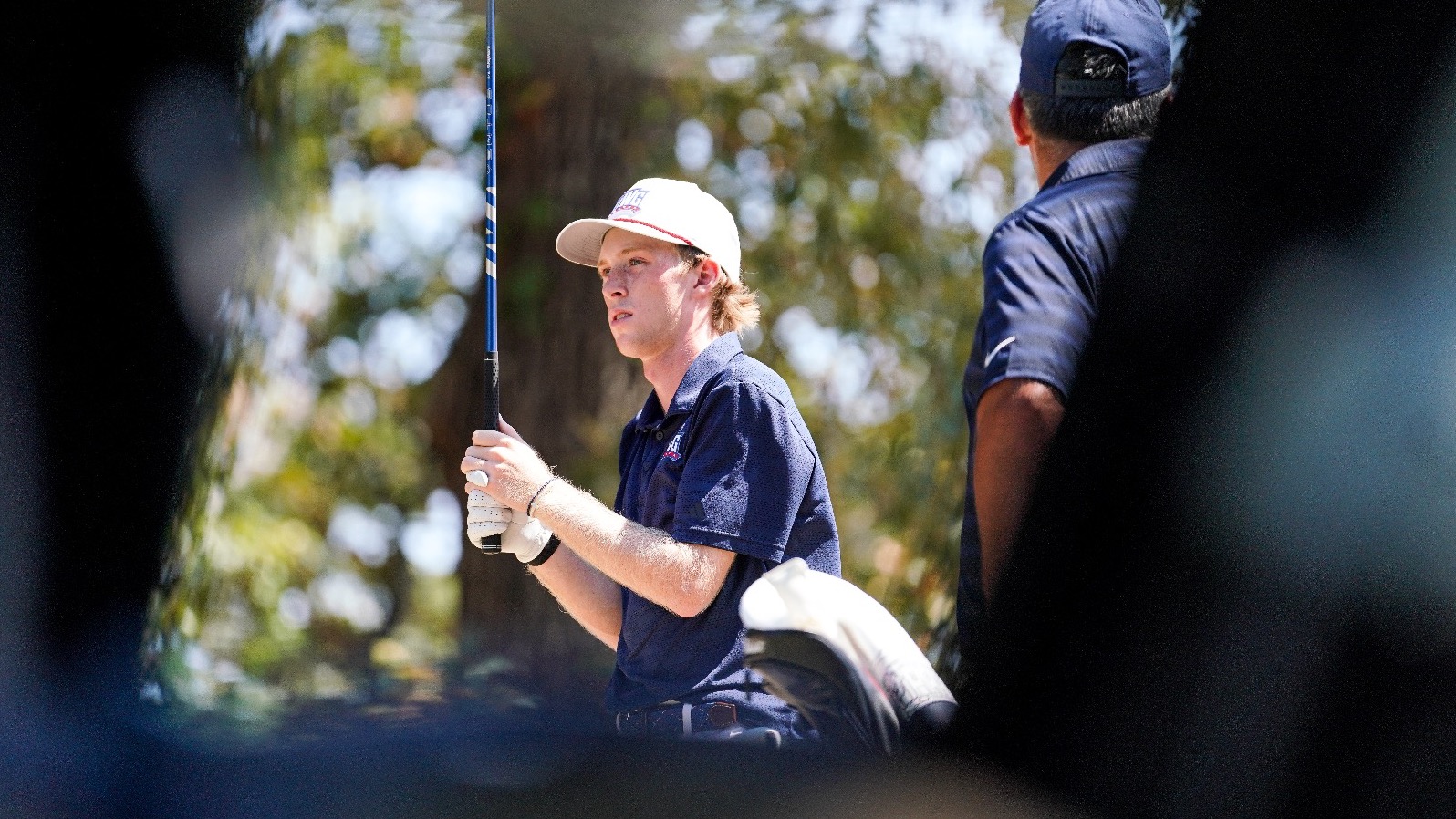 Reid Rockett looks down the fairway at a golf tournament. He is framed by a golf cart out of focus.