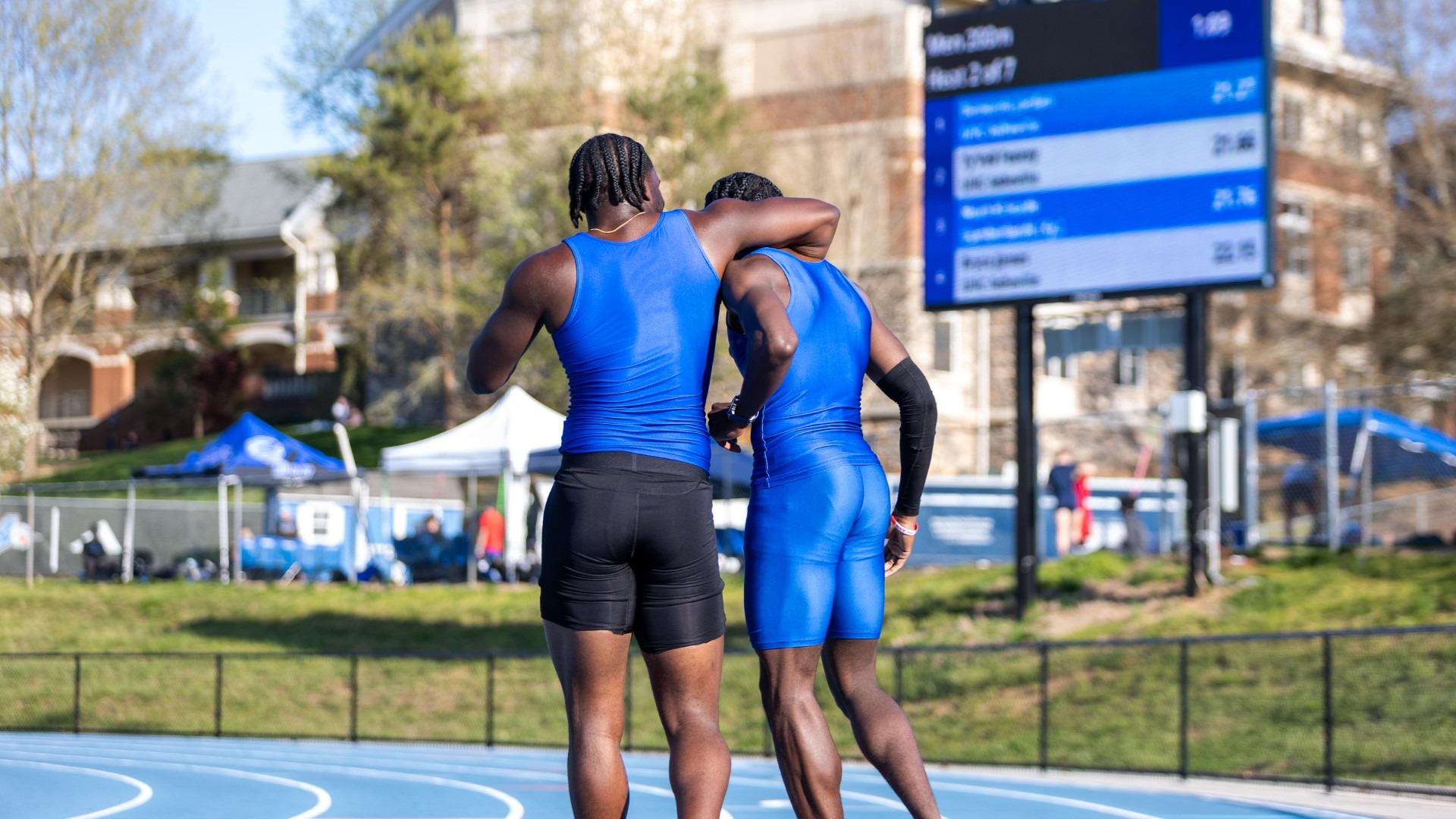 Two track athletes in blue uniforms walk together on an outdoor track, one with an arm around the other’s shoulders, with a scoreboard and campus buildings in the background.