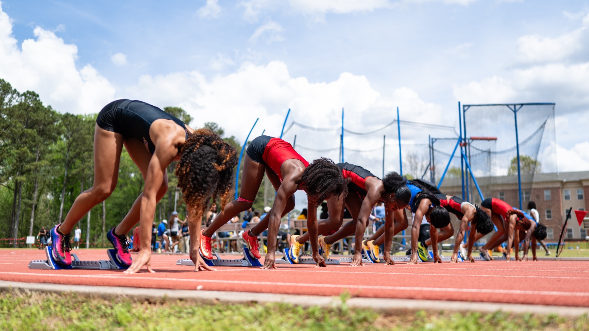 Female sprinters crouched in starting blocks on an outdoor track, preparing to begin a race under a bright sky, with spectators and field equipment visible in the background.