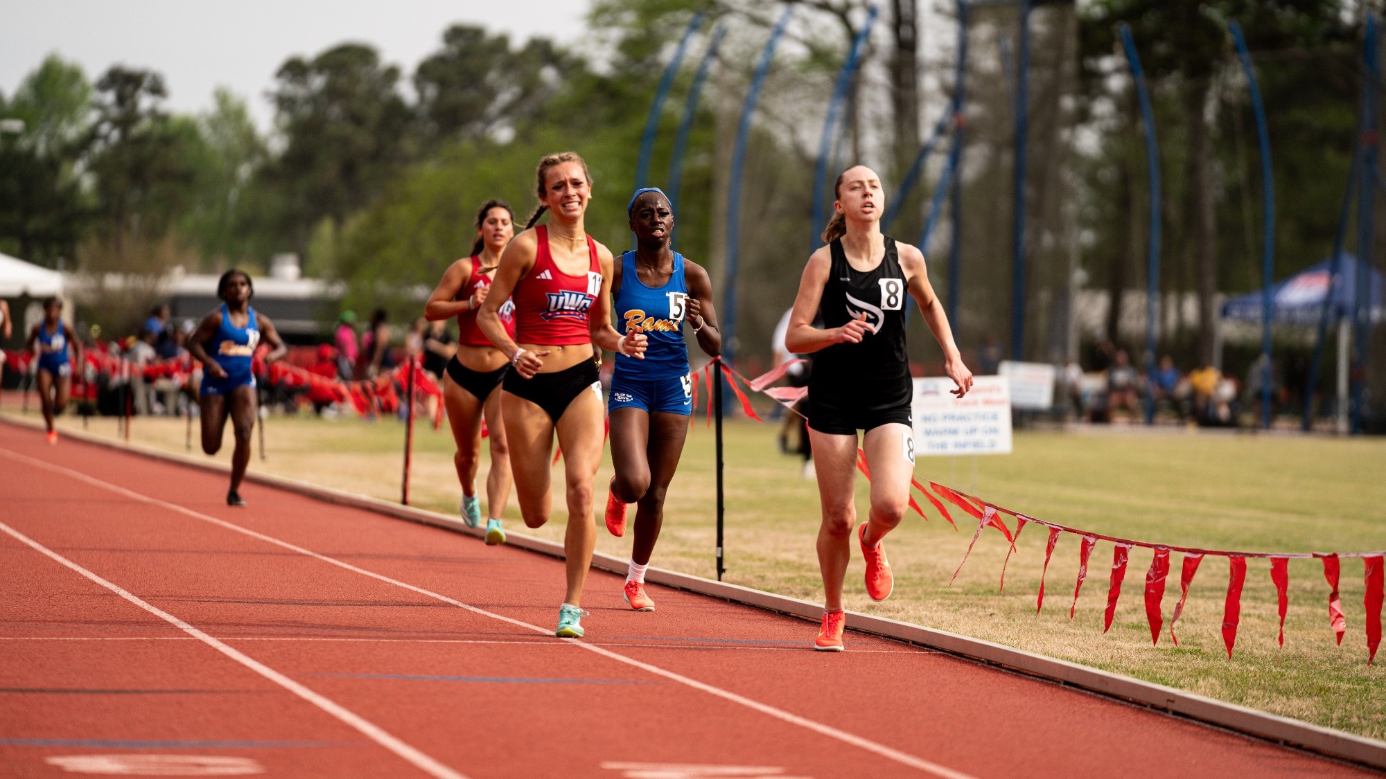 Female track athletes sprint down a red outdoor track during a race, with two runners in the lead, one in a black uniform and one in a red uniform—closely followed by competitors in blue and red kits. Spectators and other athletes are visible in the background near the infield, with trees and field equipment beyond the track.