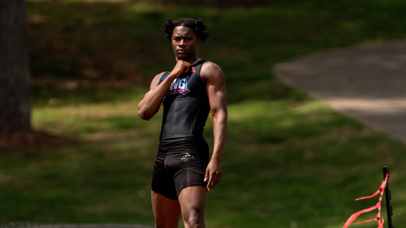 Male track athlete in a black uniform stands at the end of a runway near the track, adjusting his collar and looking focused, with cones and red boundary flags nearby and a grassy, tree-lined background behind him.