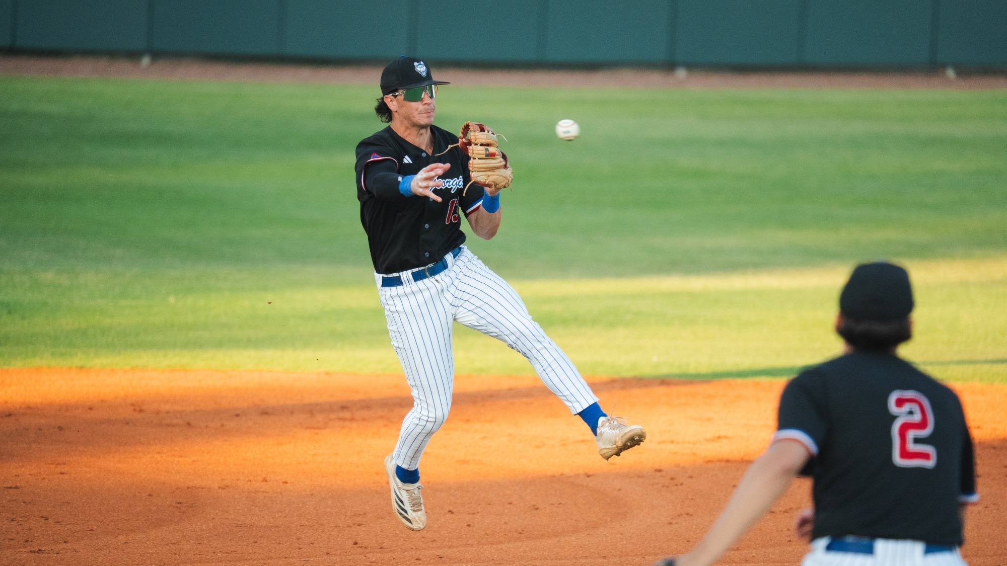 UWG baseball player in black with sun glasses and hat jumping and throwing baseball towards first base