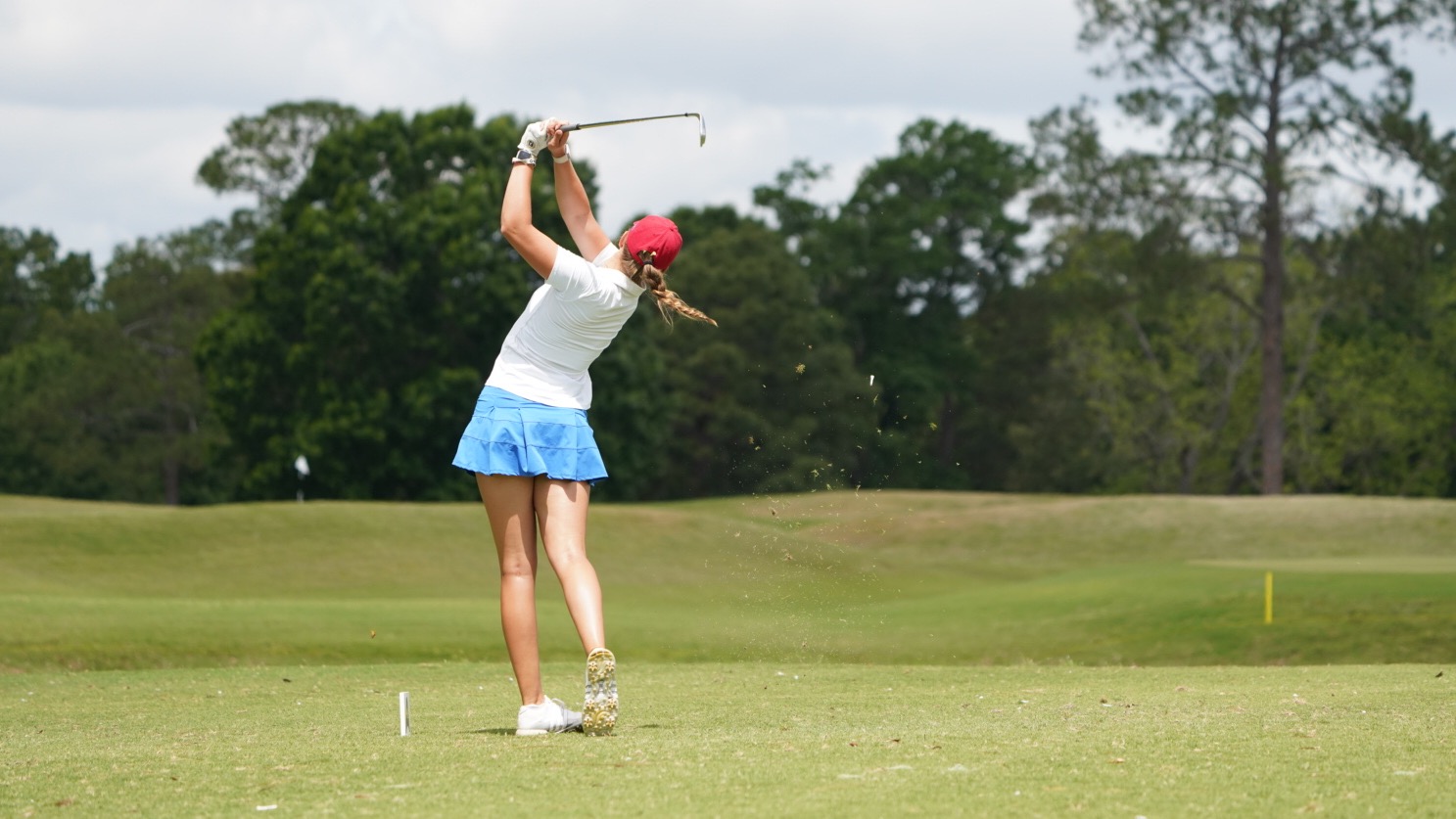 UWG women's golfer Keira Wolf hits an iron shot off of the tee of the fifth hole in the second round of the ASUN women's golf championship. She is wearing a red hat, white shirt, and blue skirt.