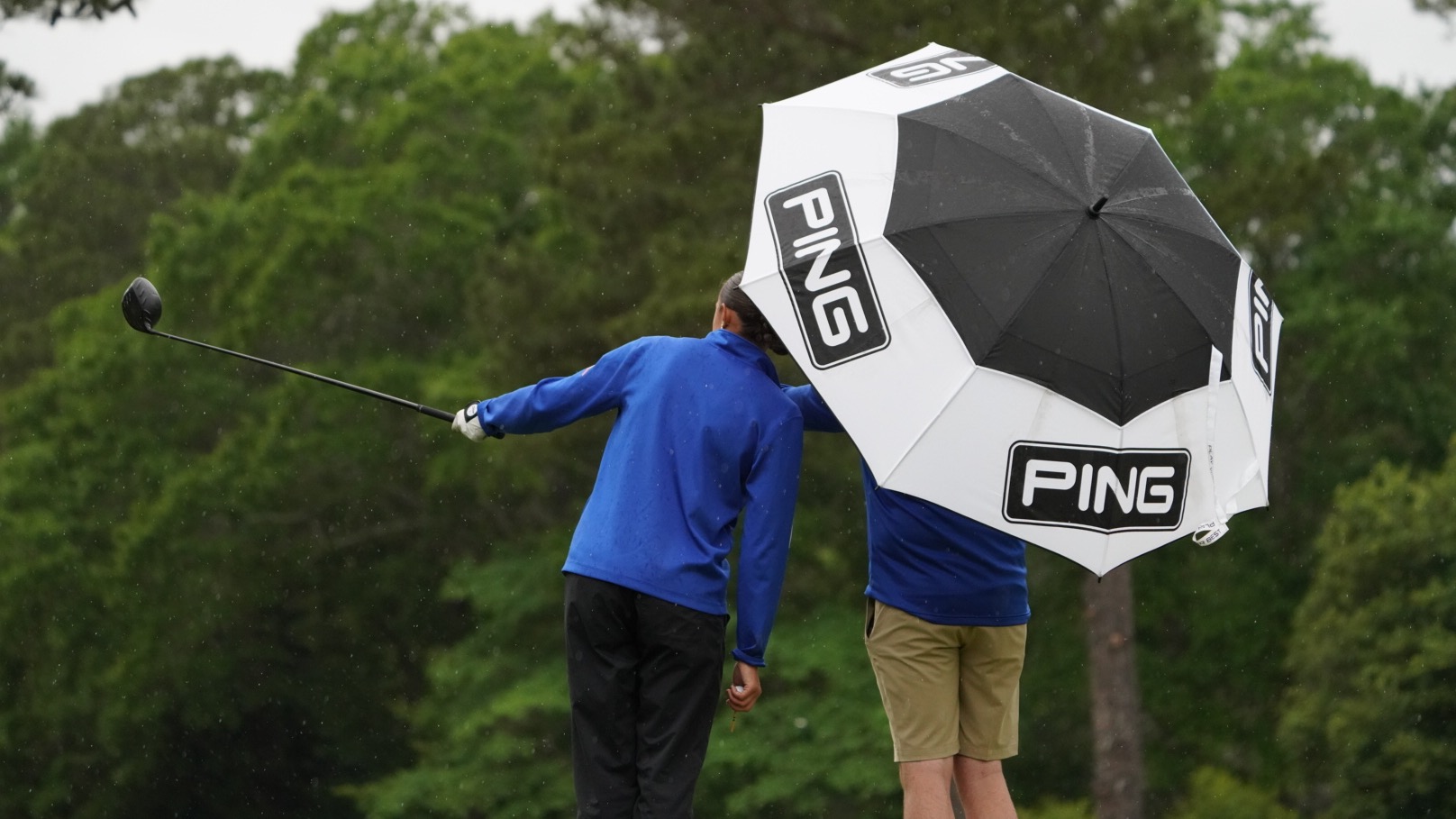 Head Coach Will Snodgrass stands under an umbrella as he and Erin Sullivan talk strategy during the third round of the ASUN Women's Golf Championships. Sullivan is pointing her driver down the fairway.