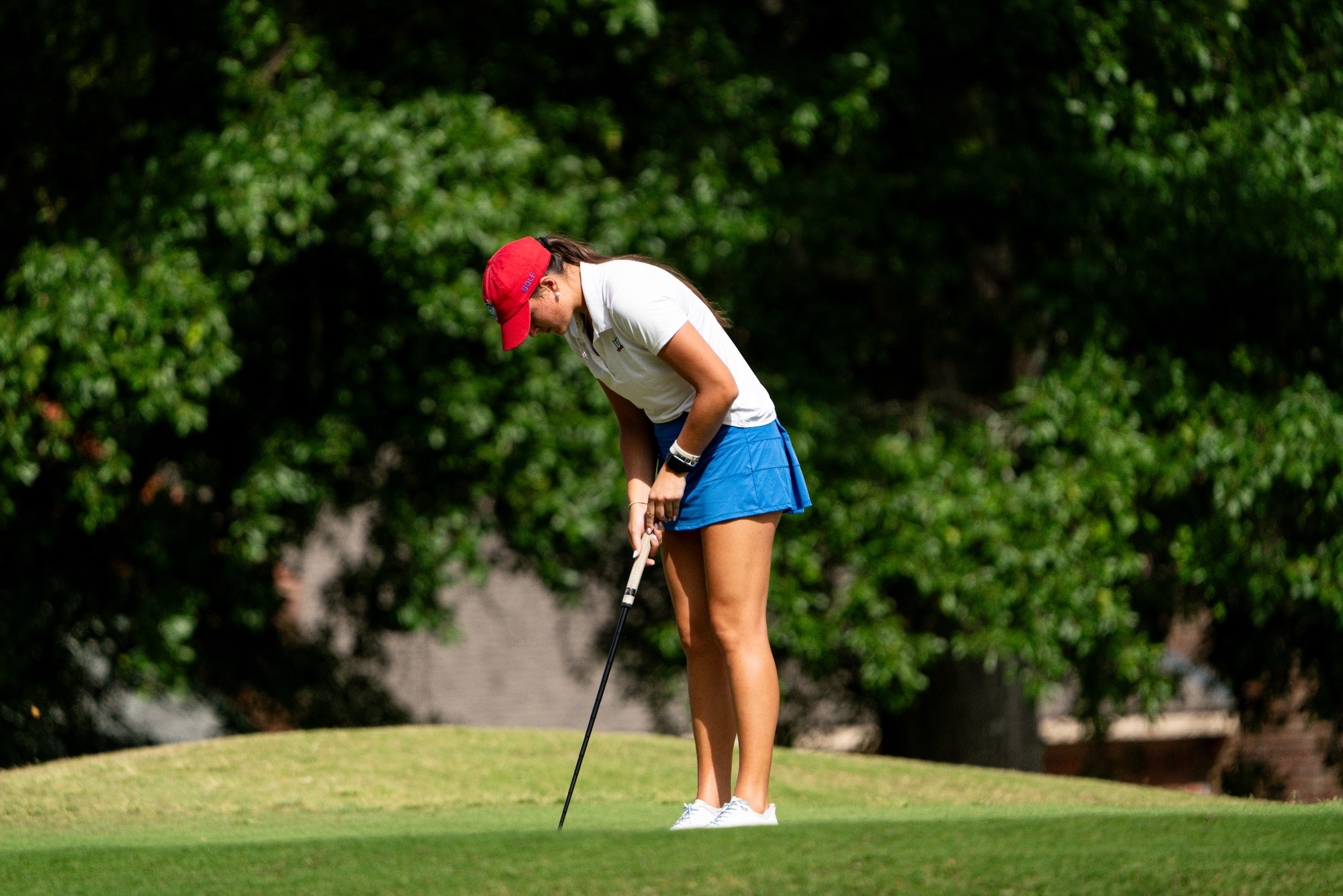 Hannah Barrett putts on the green in the second round of the ASUN Women's Golf Championships. She is wearing a red hat, white top, and blue skirt.