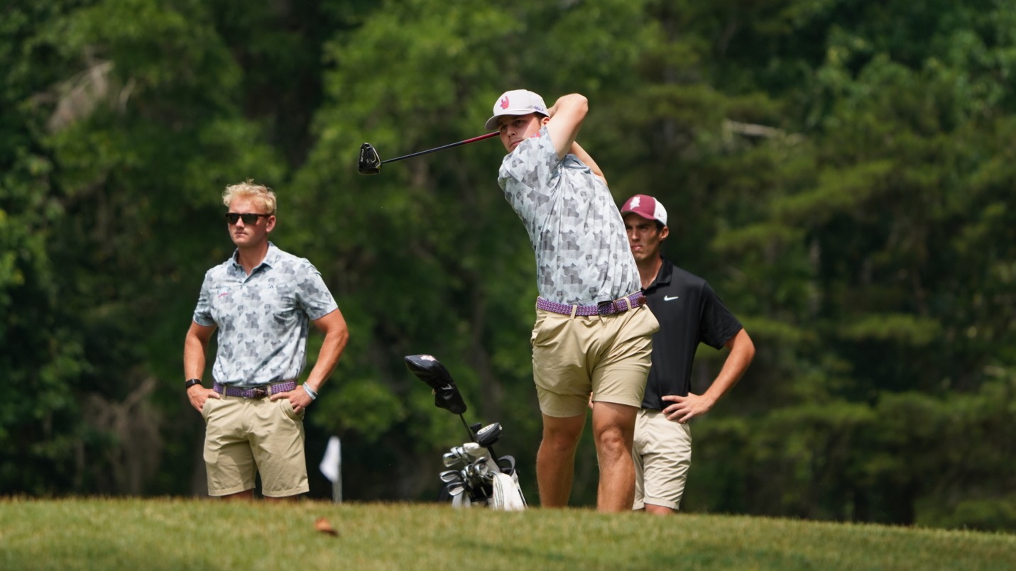 Jeremy Edmunds tees off while grad assistant coach Zack Coker looks on during the second round of the ASUN Men's Golf Championship. Coker and Edmunds are in a multicolor shirt and khaki shorts.