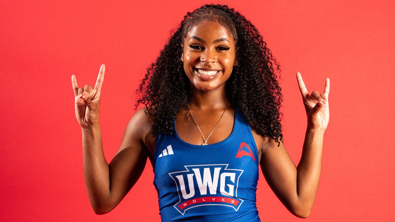 A University of West Georgia women’s track and field athlete smiles while posing in a blue UWG uniform top and black shorts against a bright red background. She raises both hands in the Wolves hand sign.