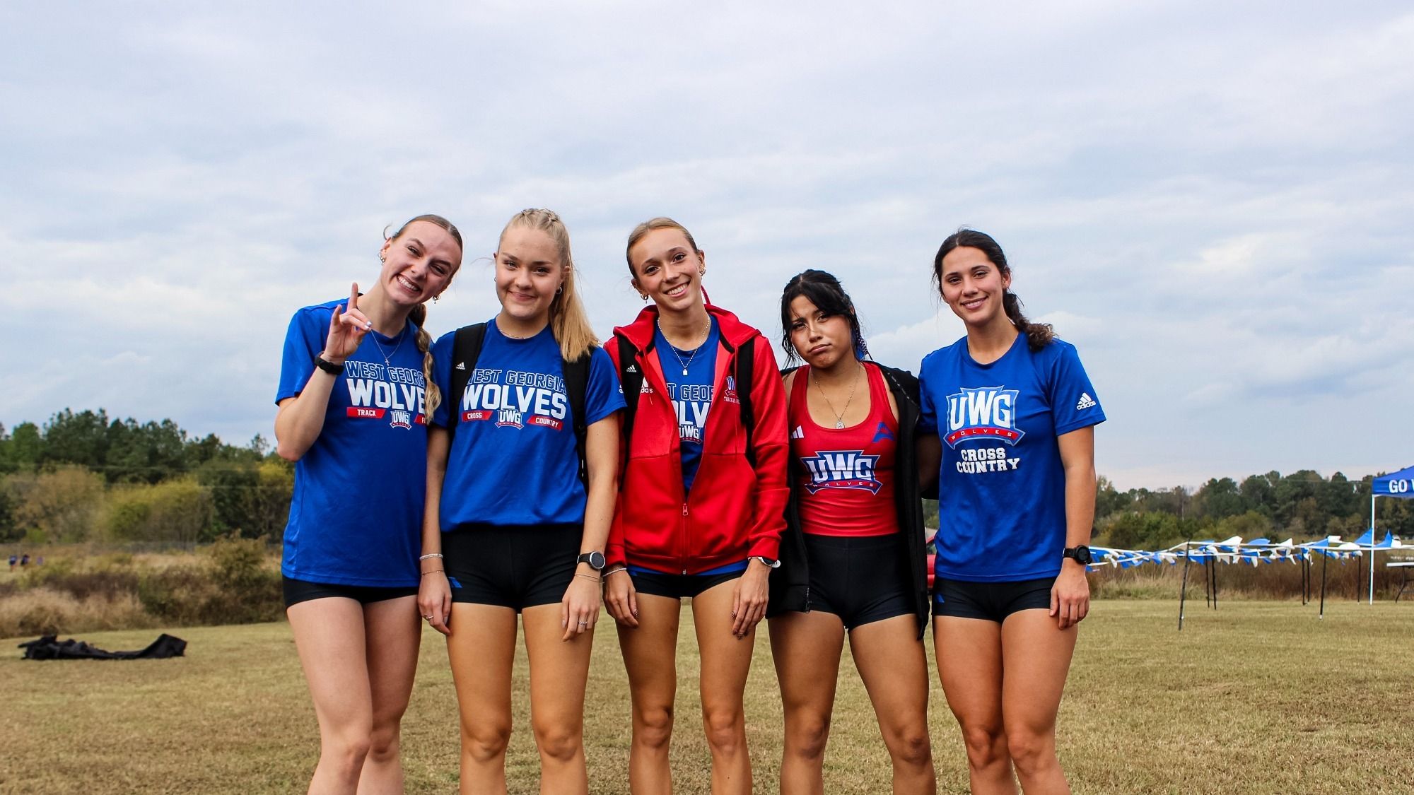 Six UWG women’s cross country athletes stand together on a grassy course, smiling and posing with arms around each other. They wear blue and red West Georgia team apparel, with race course flags and a cloudy sky in the background.