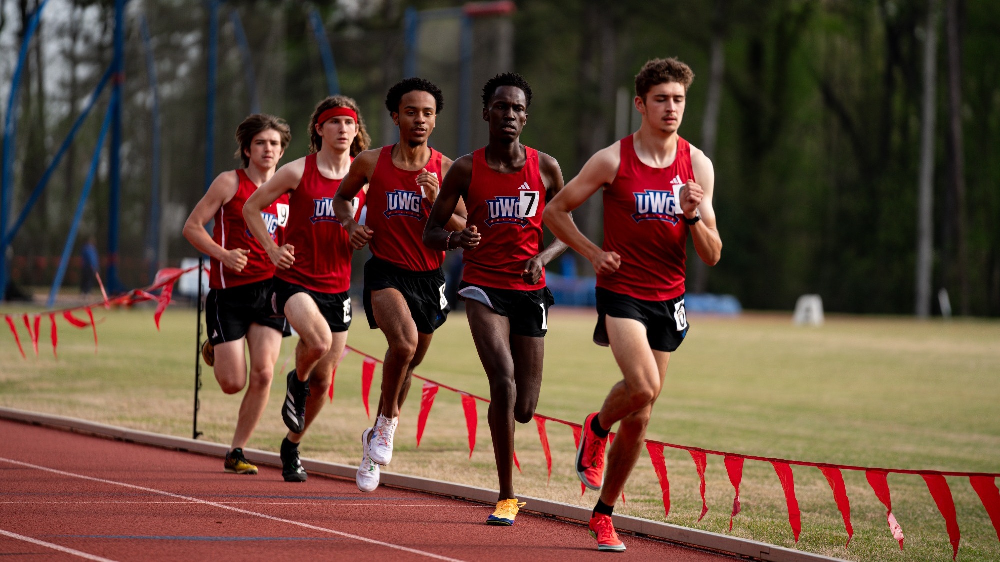 Five UWG men’s track athletes in red uniforms run together in a pack on an outdoor track, competing in a distance race with course flags lining the edge.