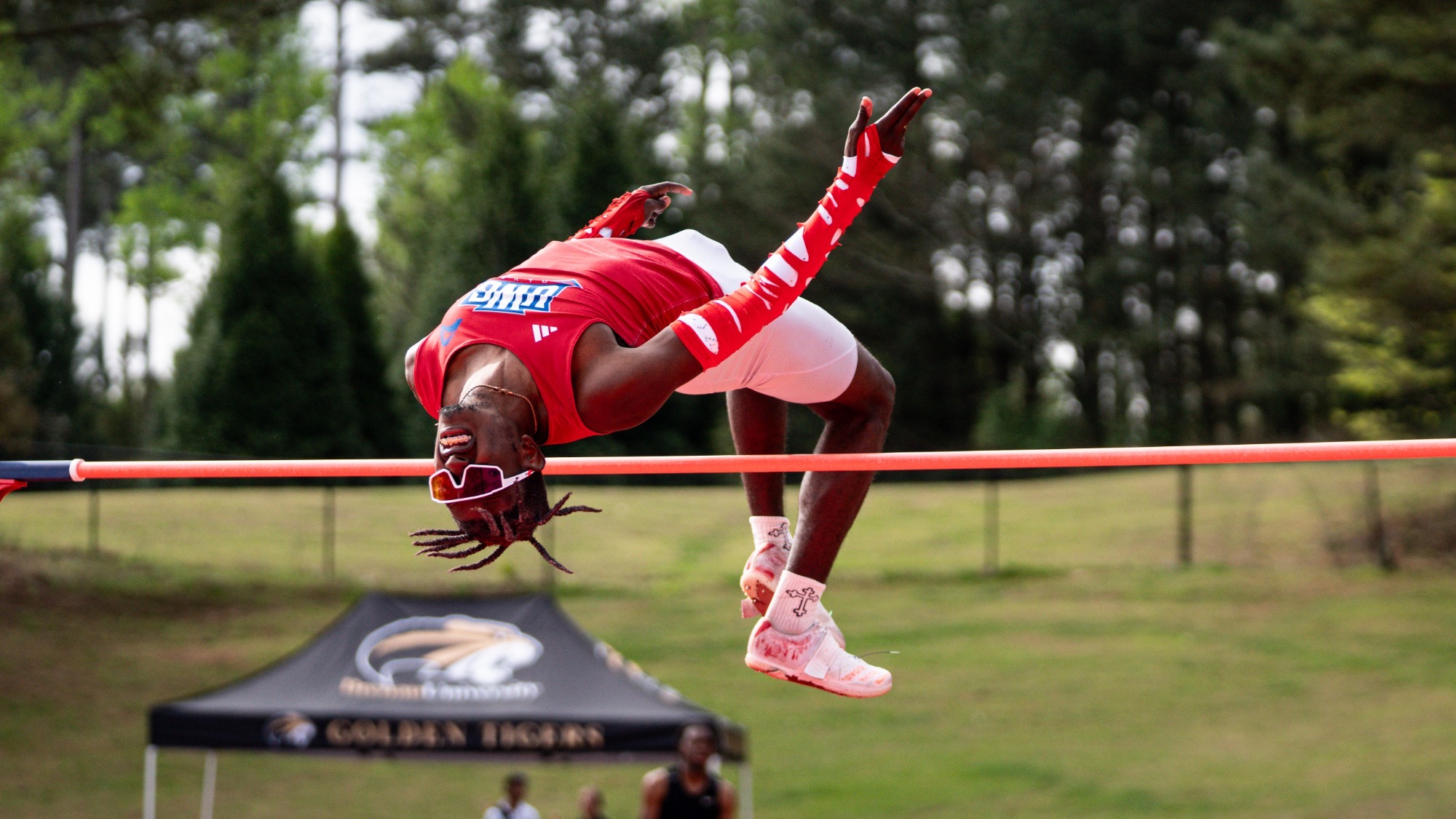 High jumper in a red uniform clears the bar mid-air using the Fosbury Flop technique, arching backward with arms extended, wearing white tights and red shoes, with a blurred outdoor track meet and spectators in the background.