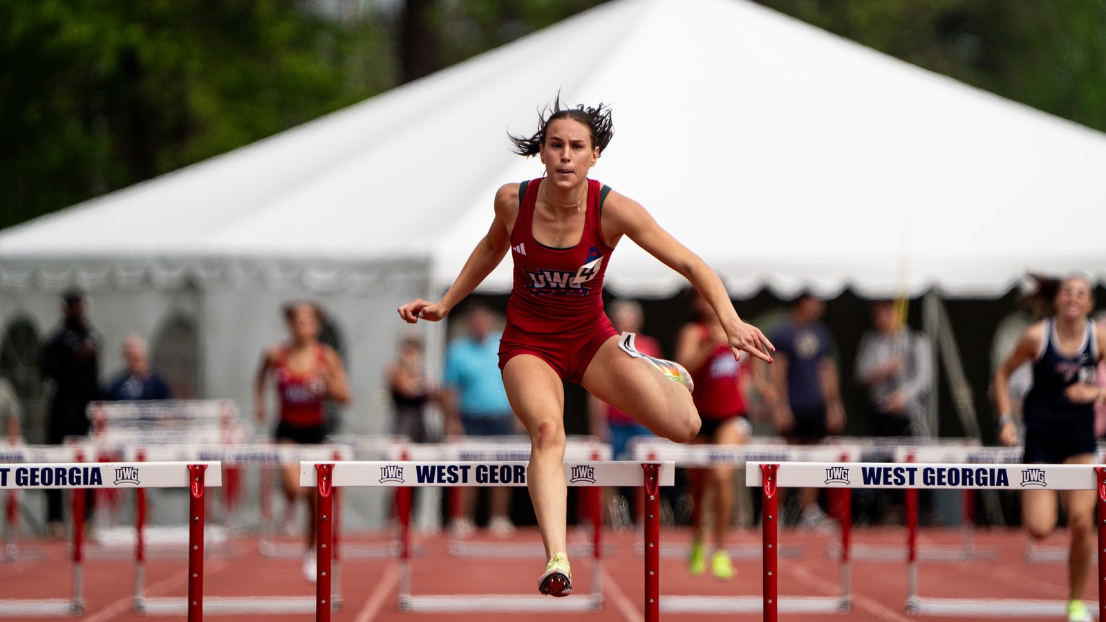 A female track athlete in a red uniform clears a hurdle during a race, with other runners and hurdles visible behind her on an outdoor track.