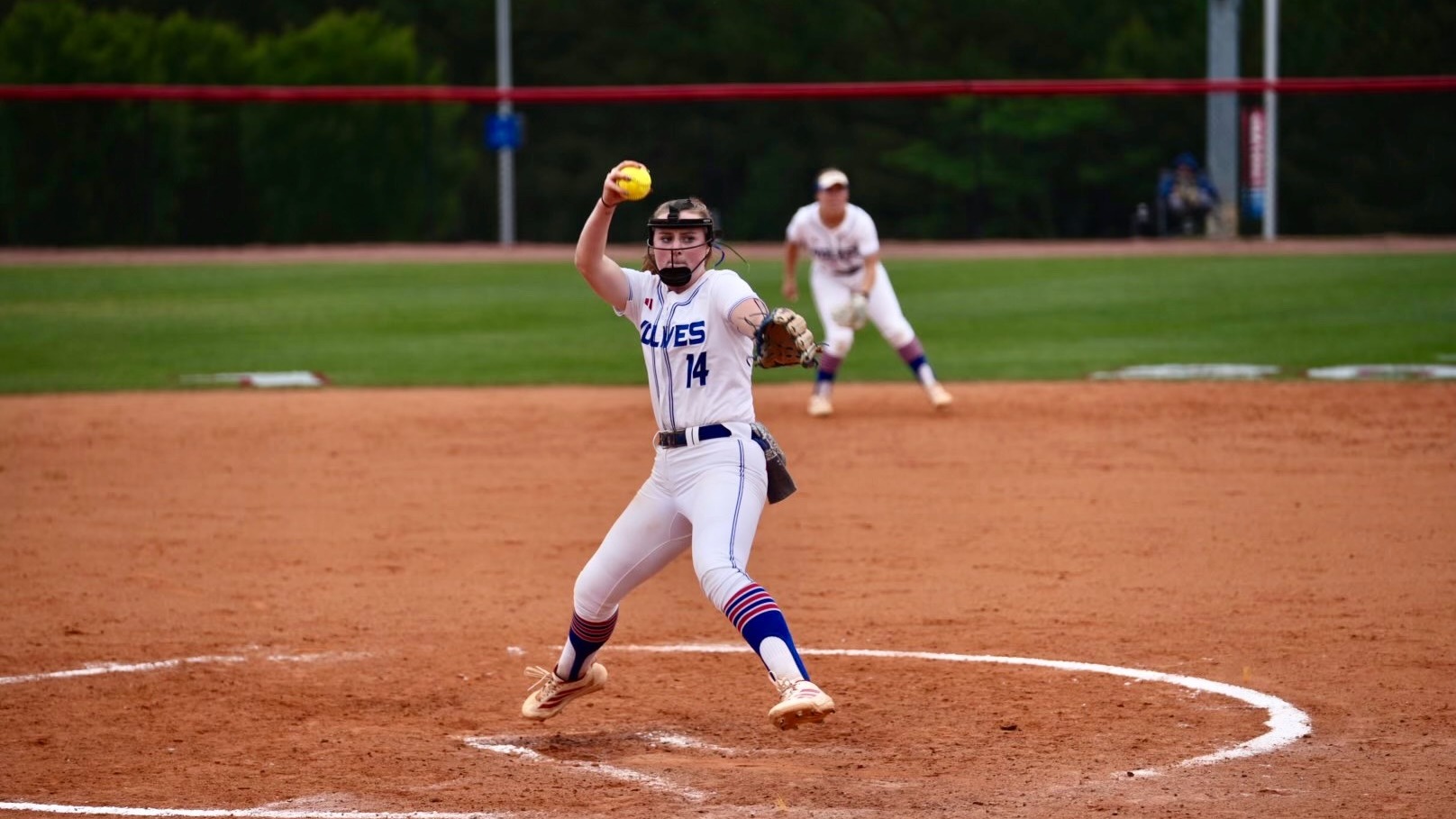 Delaney Hoyle pitches during a softball game against Queens University. She is wearing the Wolves' all white uniform. The picture is taken from the third base side of the field and the second baseman can be seen in the background.