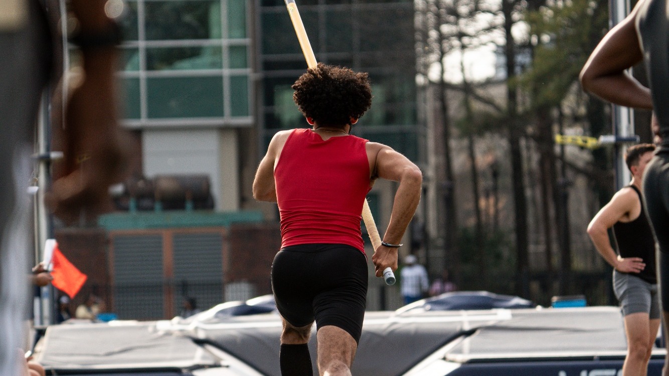 Pole vaulter in a red top sprints down the runway carrying the pole during a track and field meet, with the landing pit and standards visible ahead.