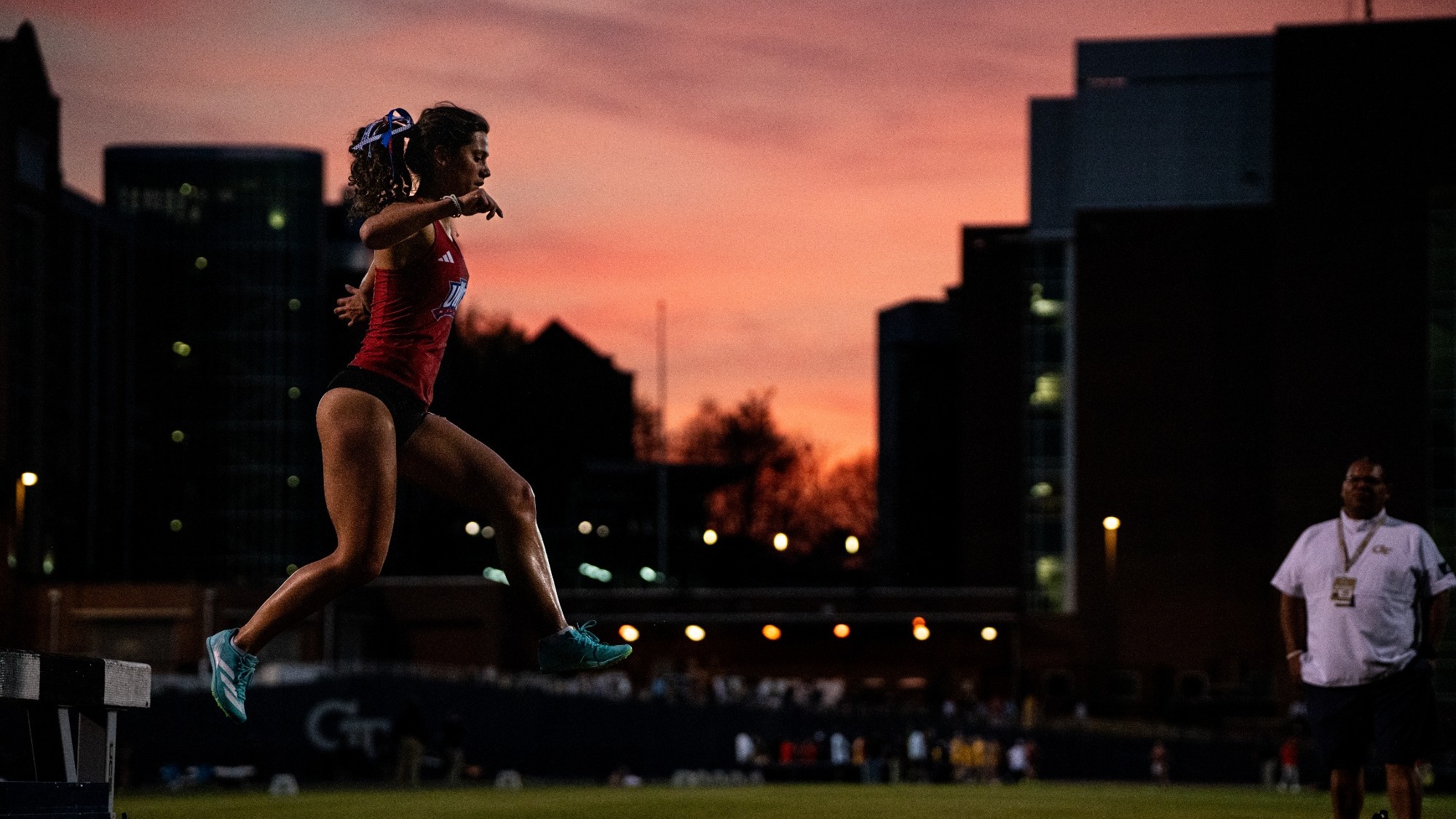 Track athlete in a red uniform leaps mid-air during a steeplechase barrier at sunset, with a city skyline and glowing lights in the background while an official watches from the side.