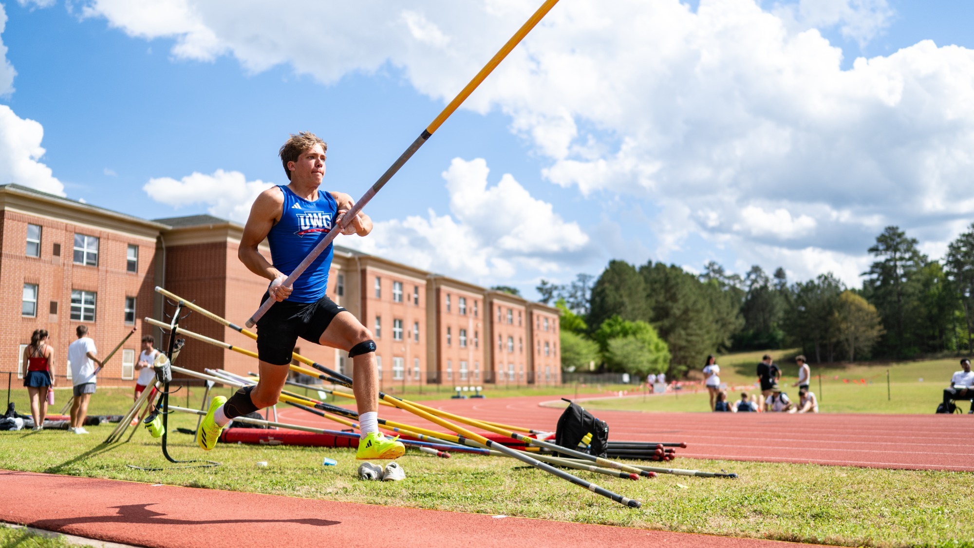 UWG pole vaulter sprints down the runway during competition at the UWG Legends Meet, carrying the pole overhead while preparing for takeoff. Multiple poles line the infield beside the track as athletes and spectators watch under a bright sky in Carrollton, Georgia.
