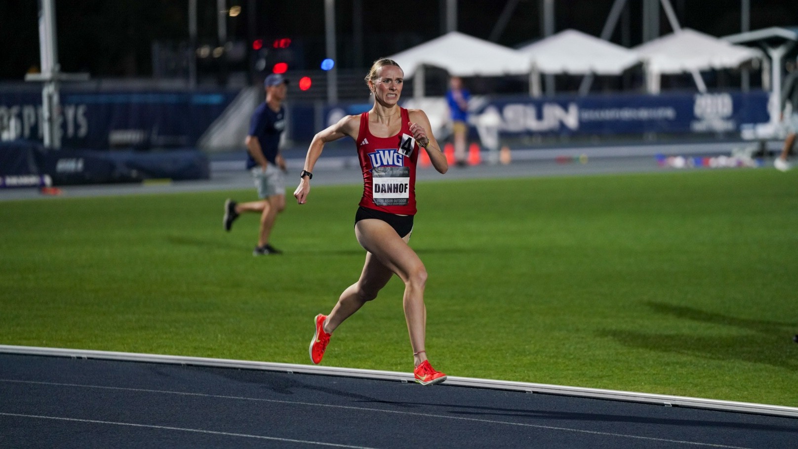 West Georgia distance runner Reatta Danhof competes during a nighttime race at the 2026 ASUN Outdoor Championships in Jacksonville, Florida. Danhof is pictured in a red UWG uniform running along the track with ASUN signage and the infield visible in the background.