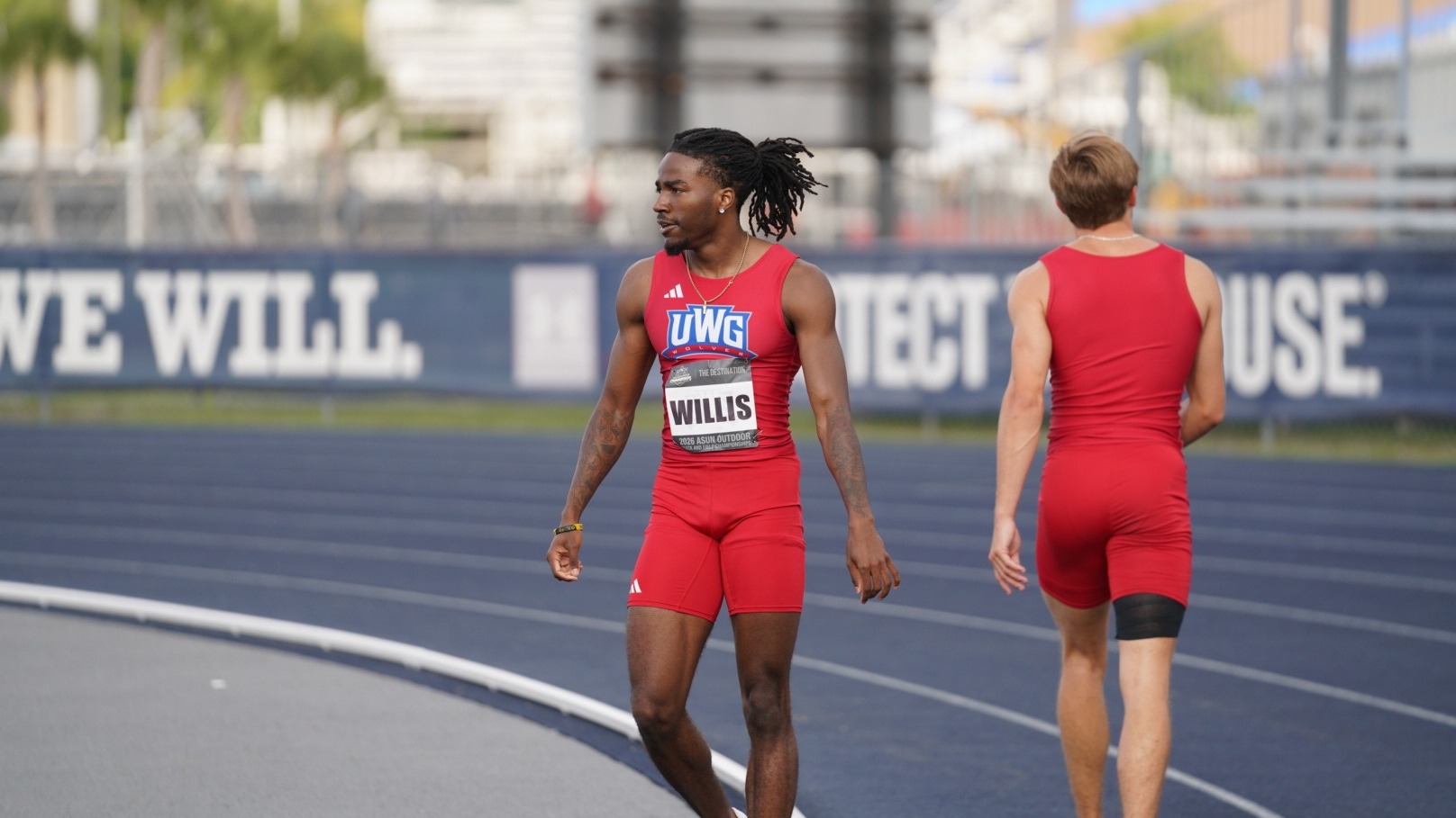West Georgia sprinter Michael Willis stands on the track during the 2026 ASUN Outdoor Championships in Jacksonville, Florida. Willis is wearing a red UWG uniform while preparing between events on the blue track surface. Another UWG athlete walks nearby in the background with stadium seating and meet signage visible behind them.